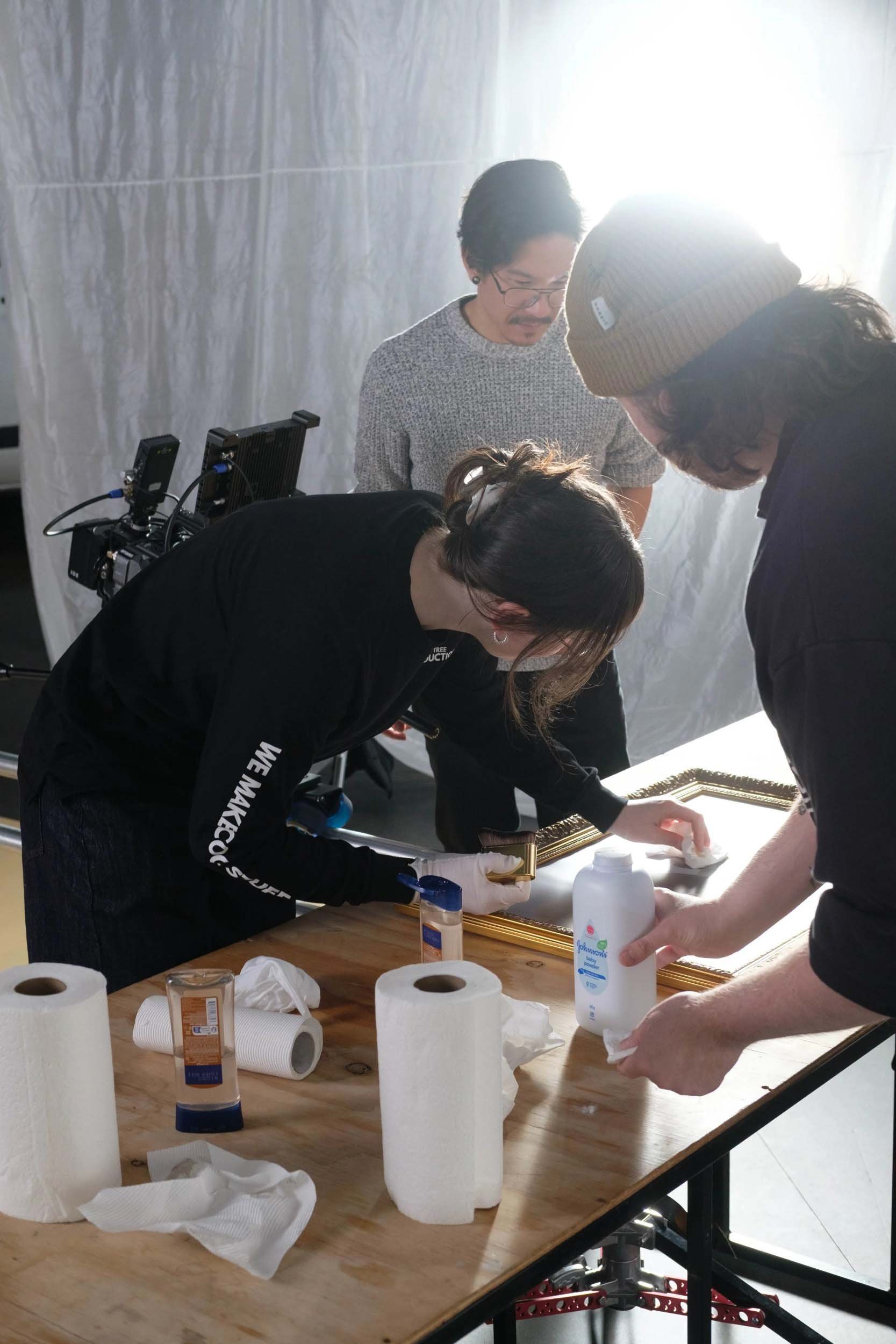 People working together on art restoration or framing project, surrounded by supplies like paper towels, glue, and picture frames, in a well-lit room.