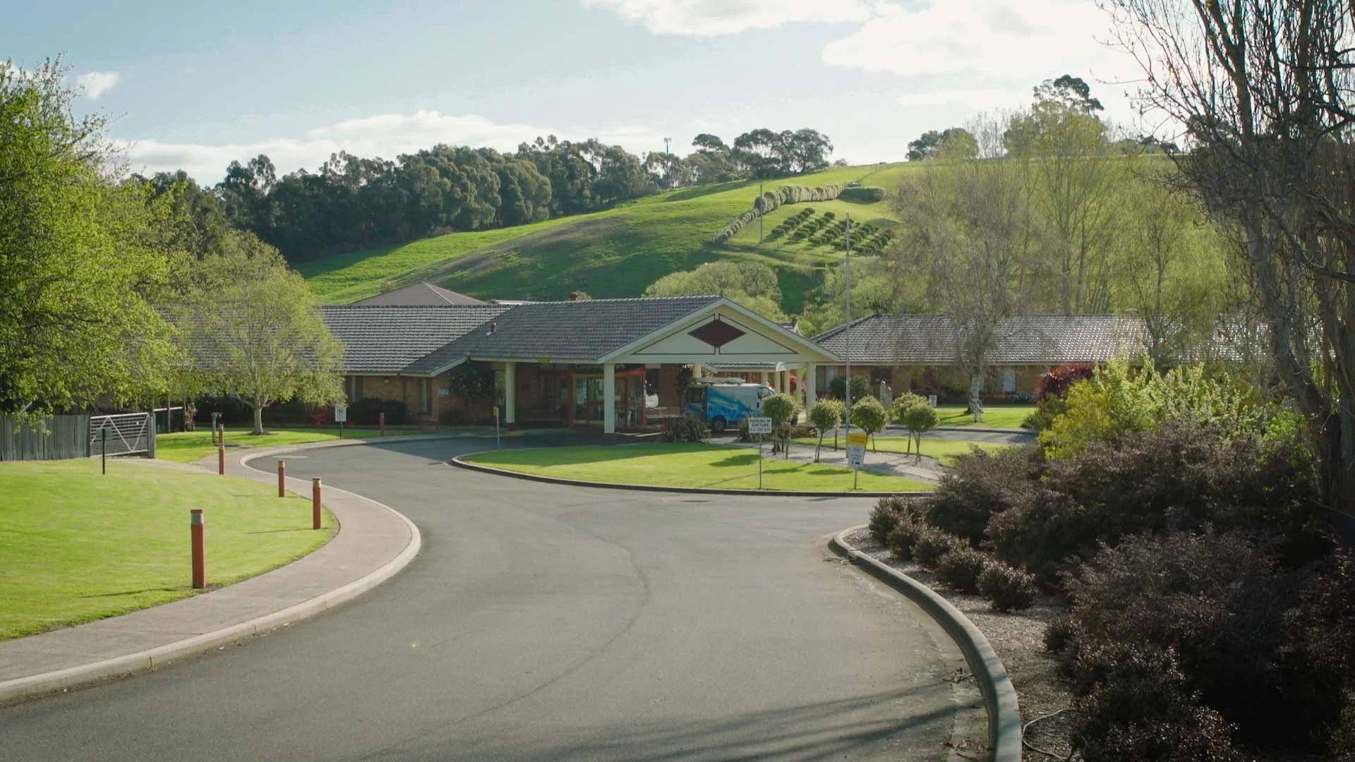 A curved driveway leading to a building with a covered entrance, surrounded by green lawns, bushes, and trees, with rolling hills and trees in the background under a partly cloudy sky.