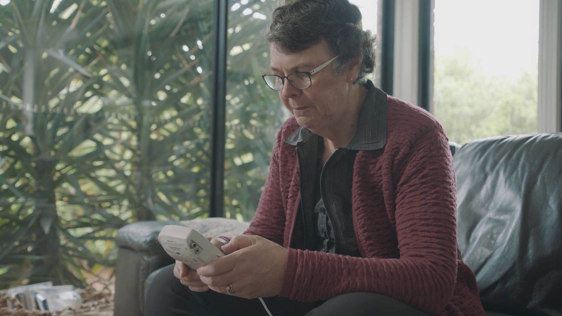 An elderly woman wearing glasses and a maroon jacket sitting on a black leather couch, looking at a remote control or medical device in her hand inside a sunlit room with large windows and greenery outside.