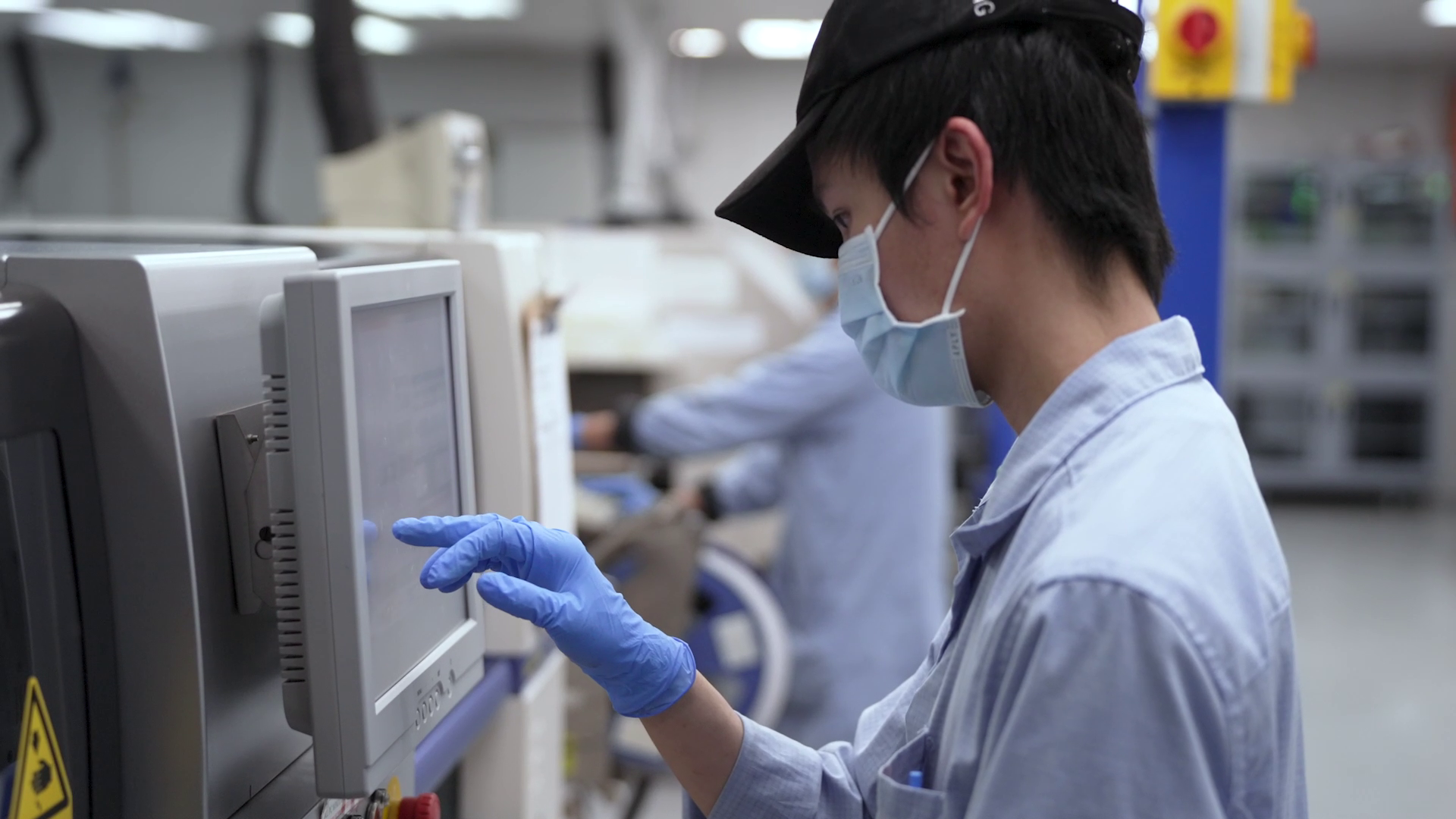 A scientist wearing a face mask, gloves, and safety glasses working with laboratory equipment, monitoring a computer screen in a lab setting.