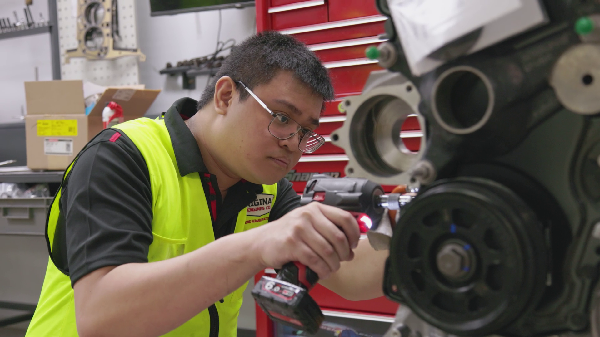 A man wearing glasses and a yellow safety vest is working on a motorcycle engine with a red cordless drill in a workshop.