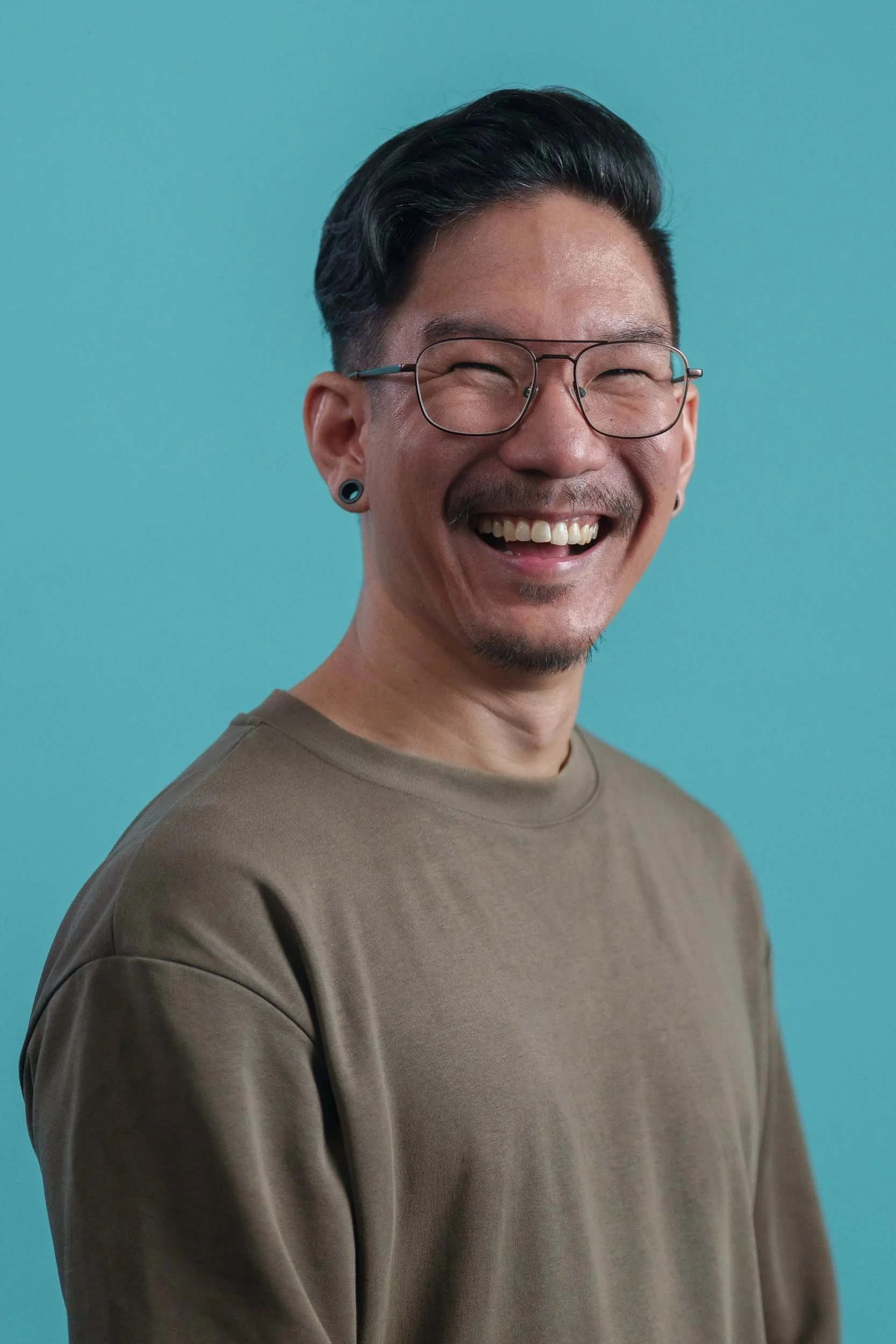 A man with black hair, glasses, earrings, and a goatee, smiling broadly against a teal background, wearing a brown shirt.