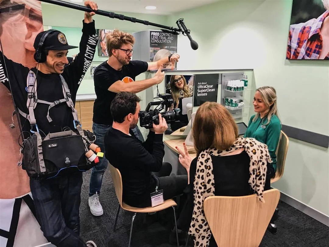 Matt Smolen using his hands to frame up the shot whilst, Tim holds the boom over head and Dan sits in a chair filming a Specsavers team member.