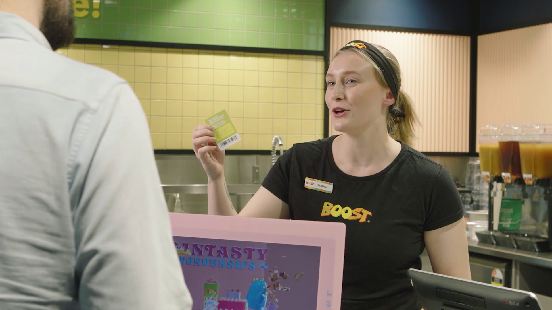A Boost employee with a name tag interacting with a male customer at a smoothie store counter. The employee is holding a green card and appears to be explaining something.