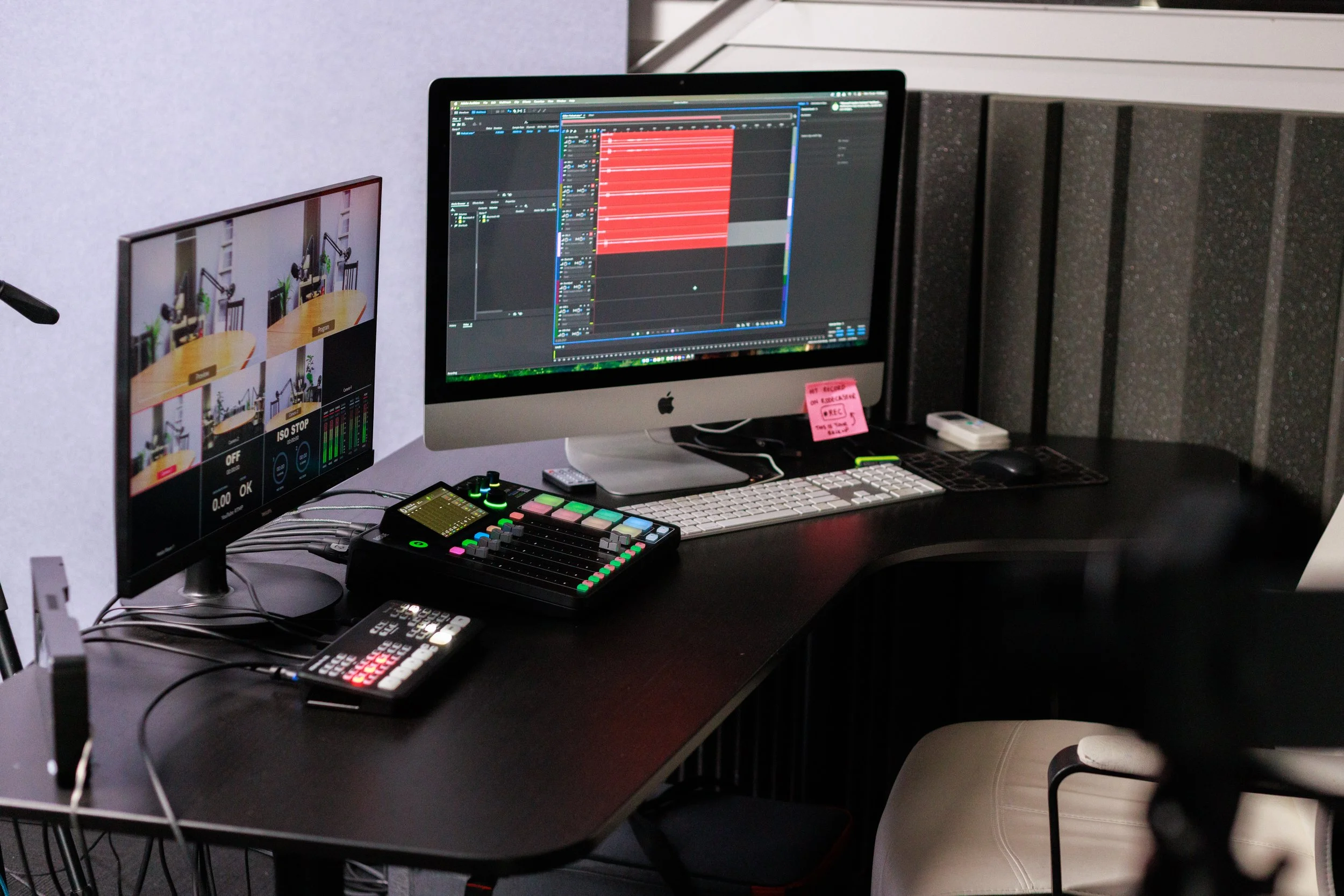 A production workspace with two computer monitors, one displaying video editing software, a control surface with colorful buttons, a small black mixer, a remote control, and a keyboard and mouse on a black desk. There is also a chair with a white cus