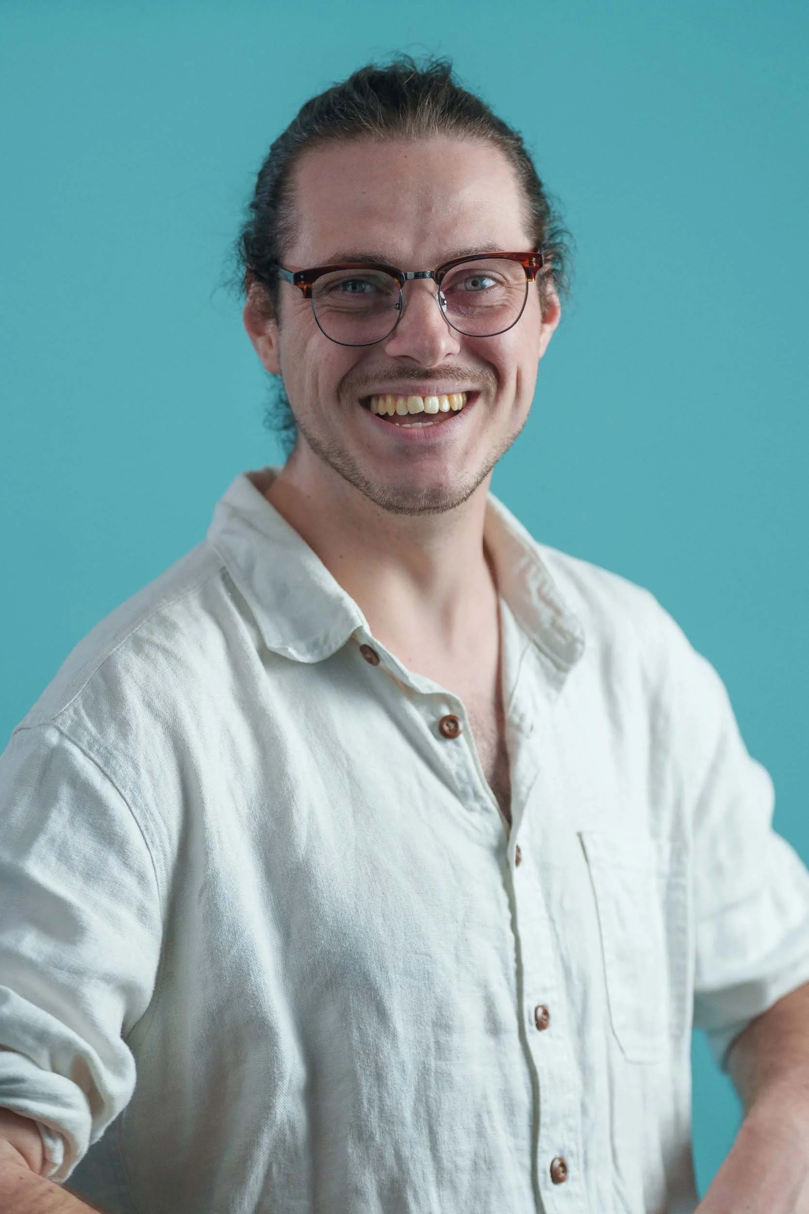 A person with glasses, smiling, wearing a light-colored button-up shirt, against a solid light blue background.
