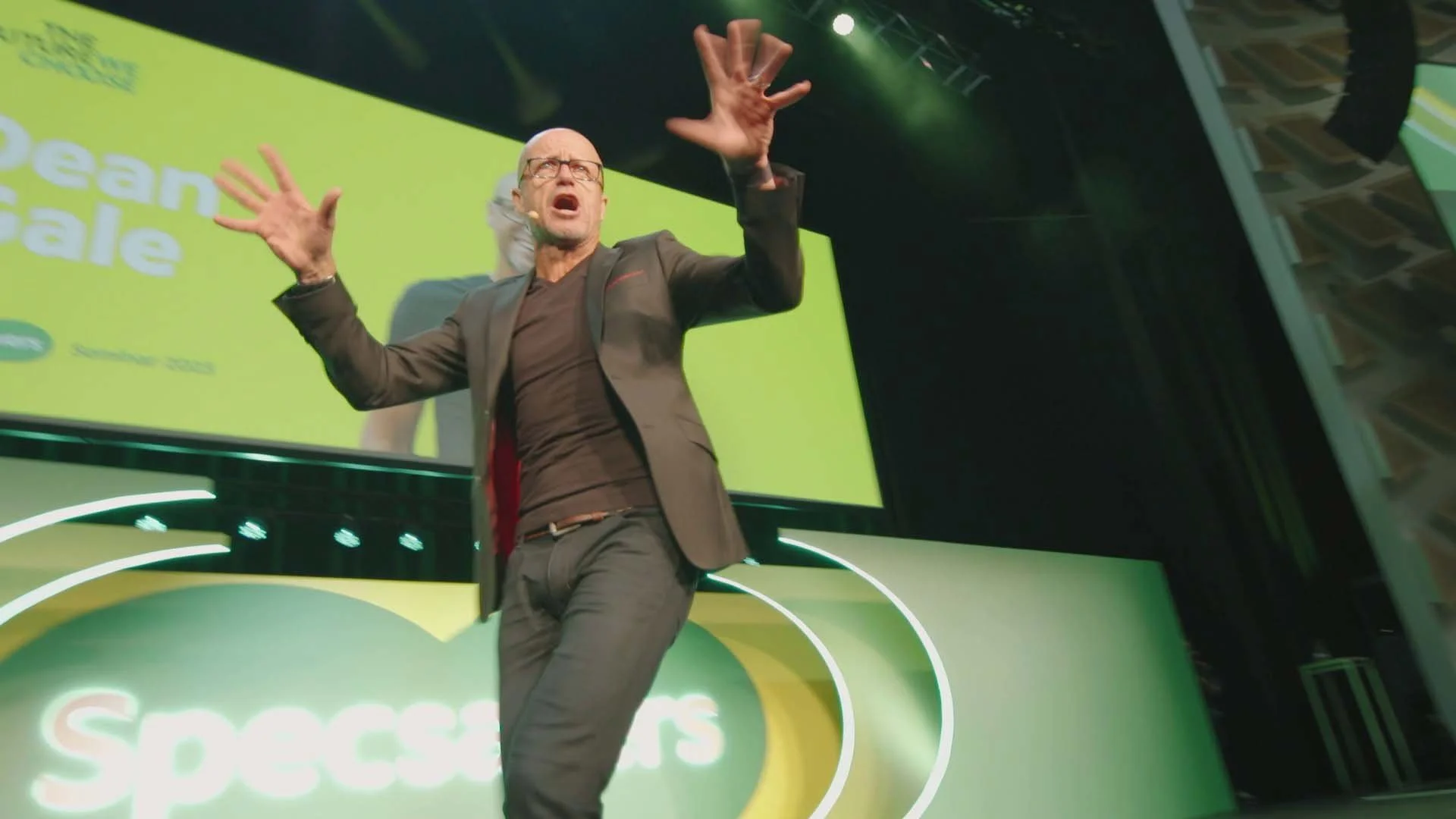A man dressed in a blazer and dark pants is standing on a stage with his arms raised and mouth open, speaking passionately. The background features a bright green screen with text and graphics, including the word 'sustainable.' The stage is decorated with green and white lighting. There is a large projection or screen behind him displaying the same vibrant background.