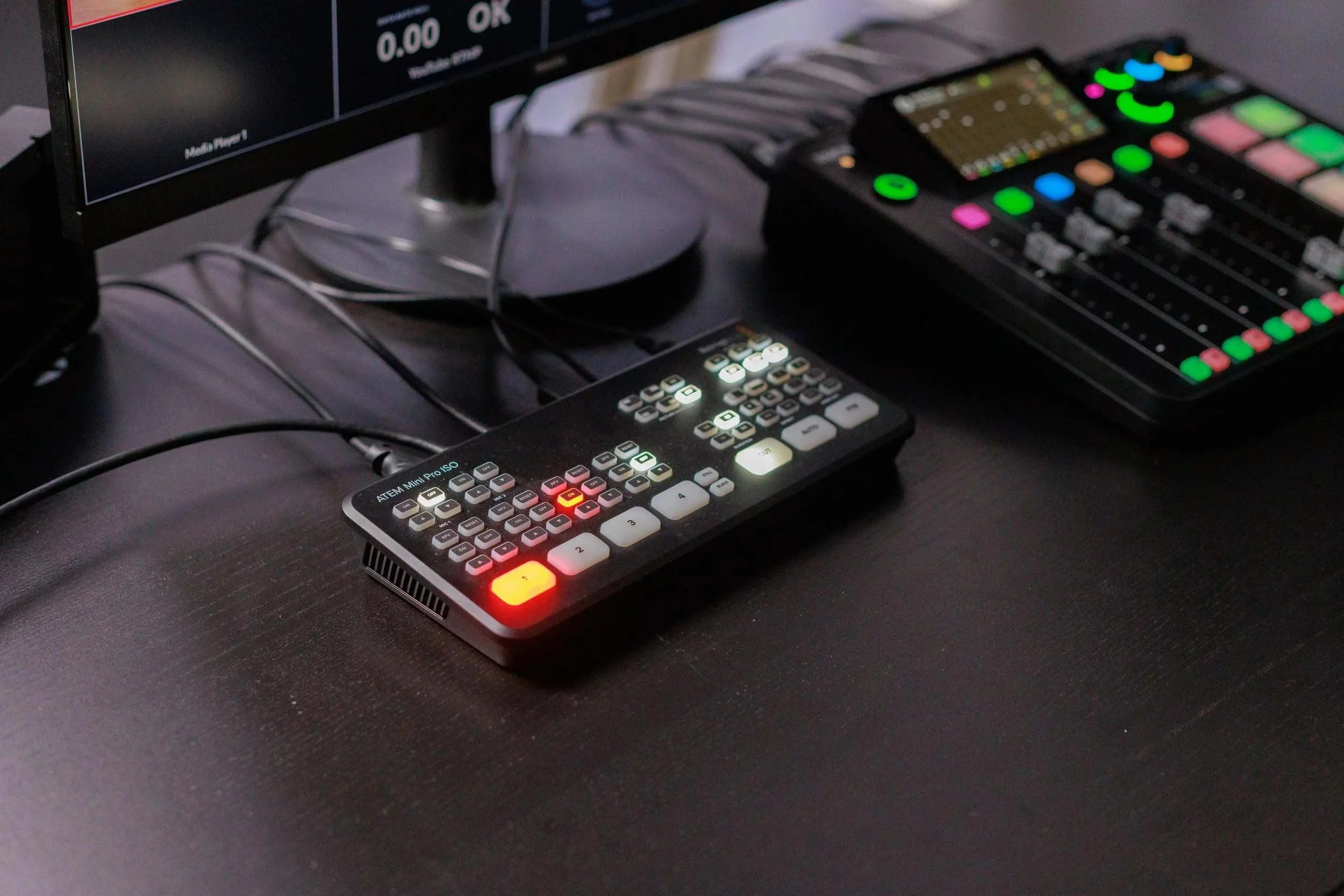 Close-up of a music production setup with a computer monitor displaying audio levels, a digital mixing console with colorful backlit pads, and a compact control device with illuminated buttons on a black surface.