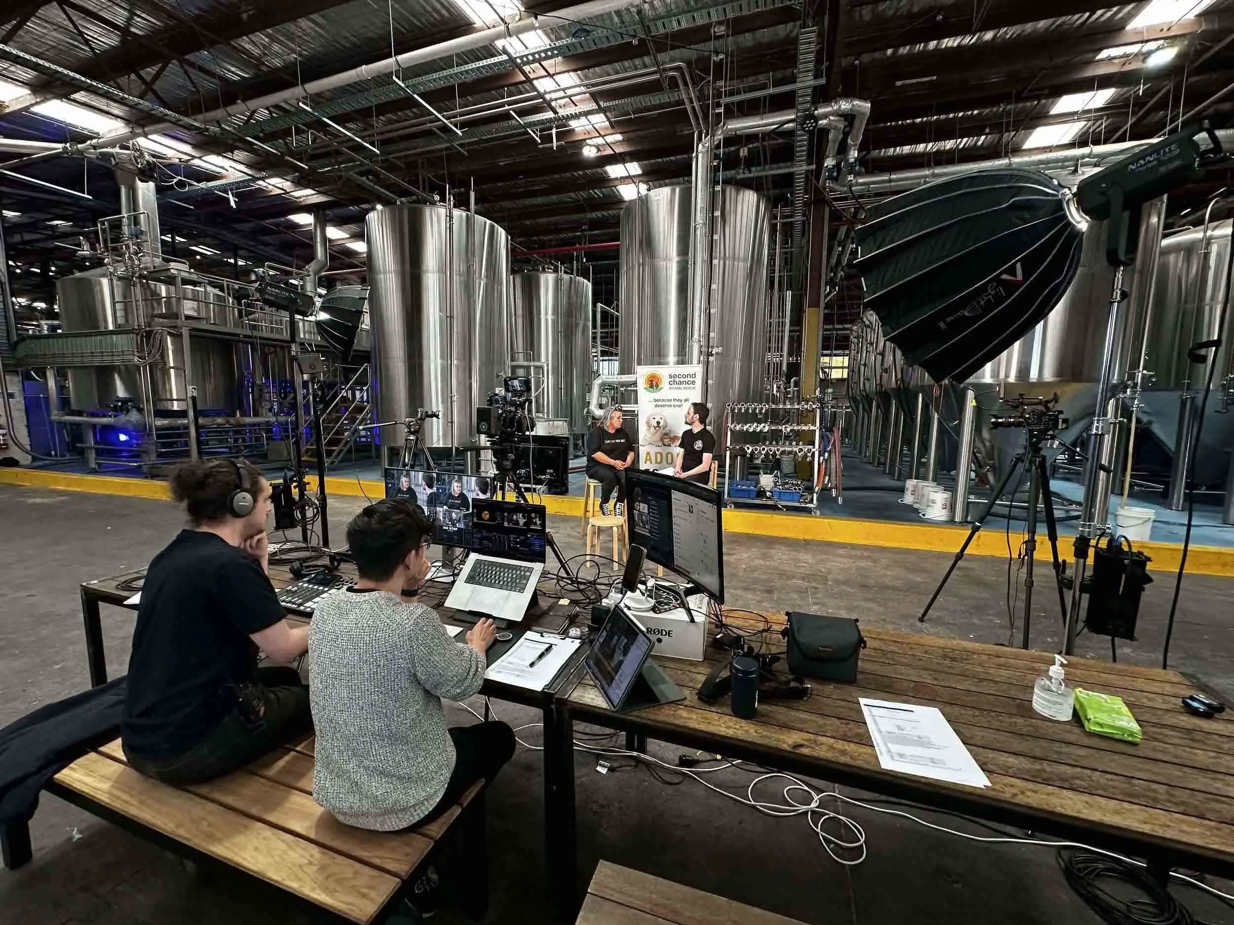 A behind-the-scenes view of a filming or interview setup at a brewery or industrial facility with large stainless steel tanks in the background. In the foreground, people are working with audio and video equipment, including cameras and laptops, while two women are seated and having a conversation near a banner that reads 'second chance animal rescue'.