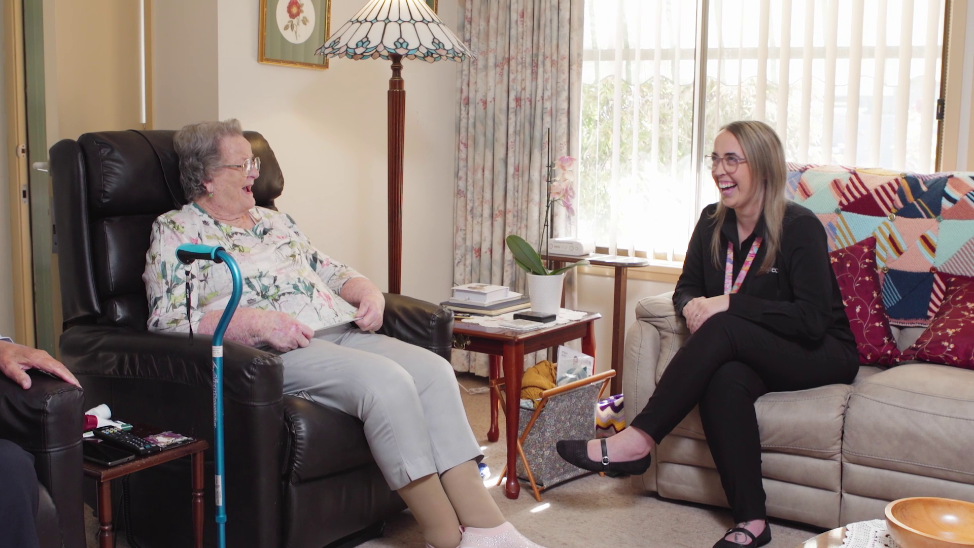 An elderly woman and a middle-aged woman are sitting and laughing in a cozy living room with a window, curtain, and various decorative items.