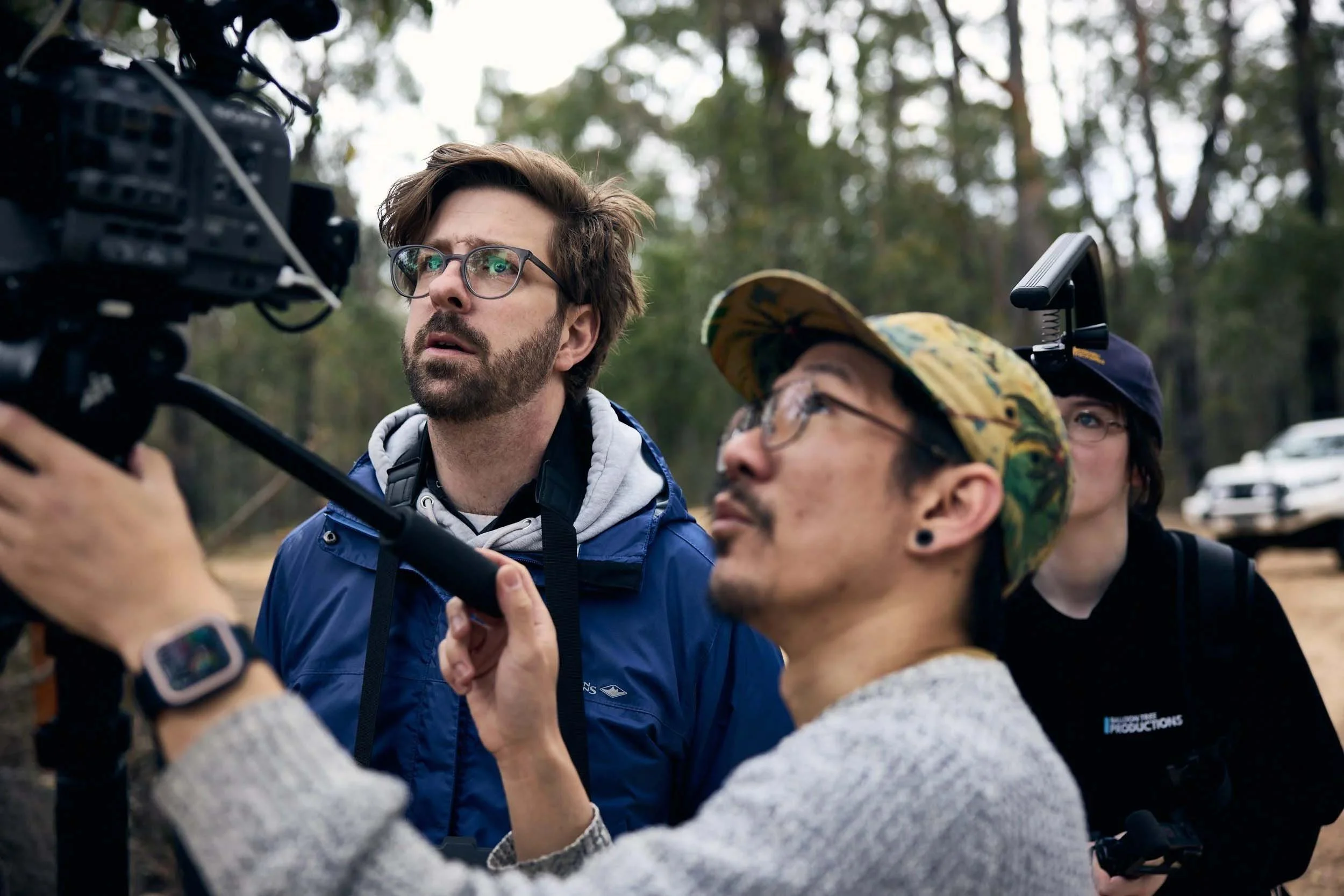 A group of three people outdoors in a forested area, gathered around a camera. One man with glasses and a beard is looking at the camera, while another woman with glasses and a cap looks on, and a third person's hands are visible near the camera.