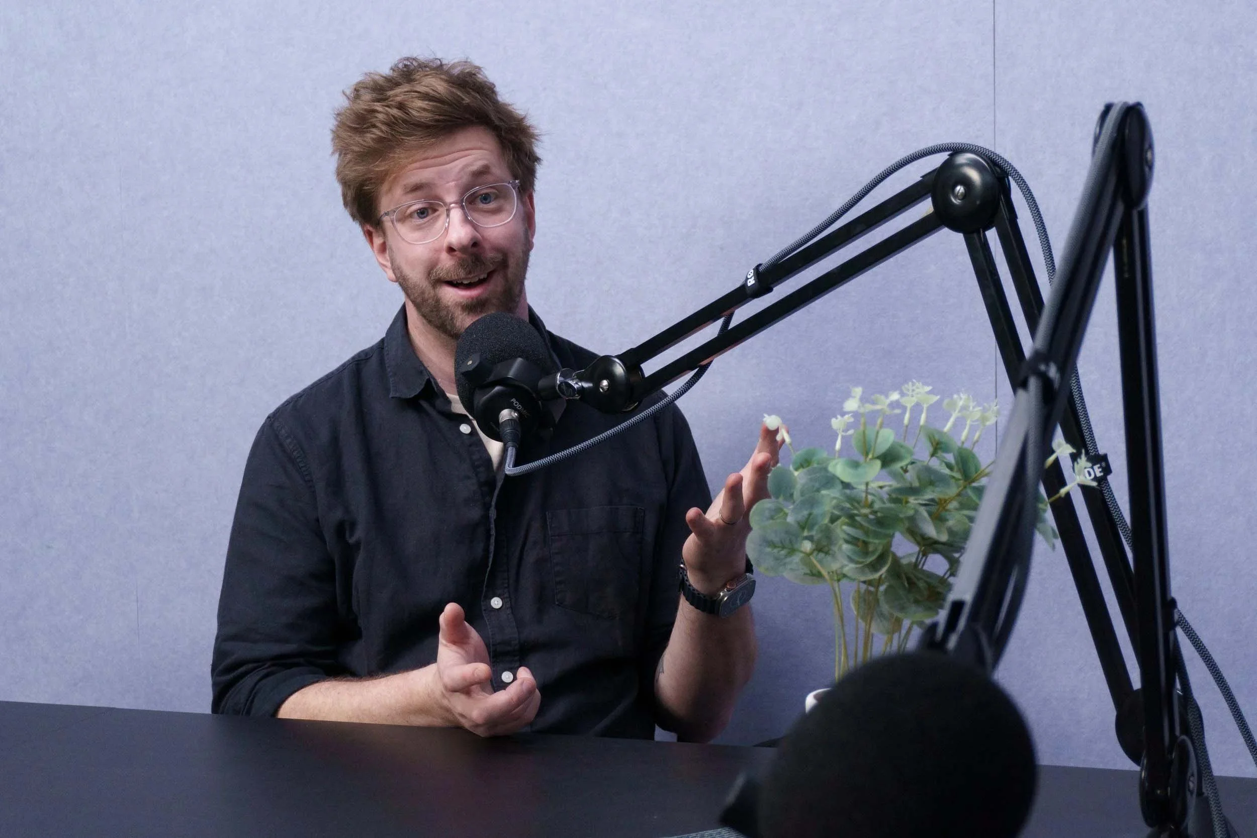 A man with glasses and a beard in a black shirt speaking into a microphone in a recording studio, with a plant on the table and a gray wall in the background.