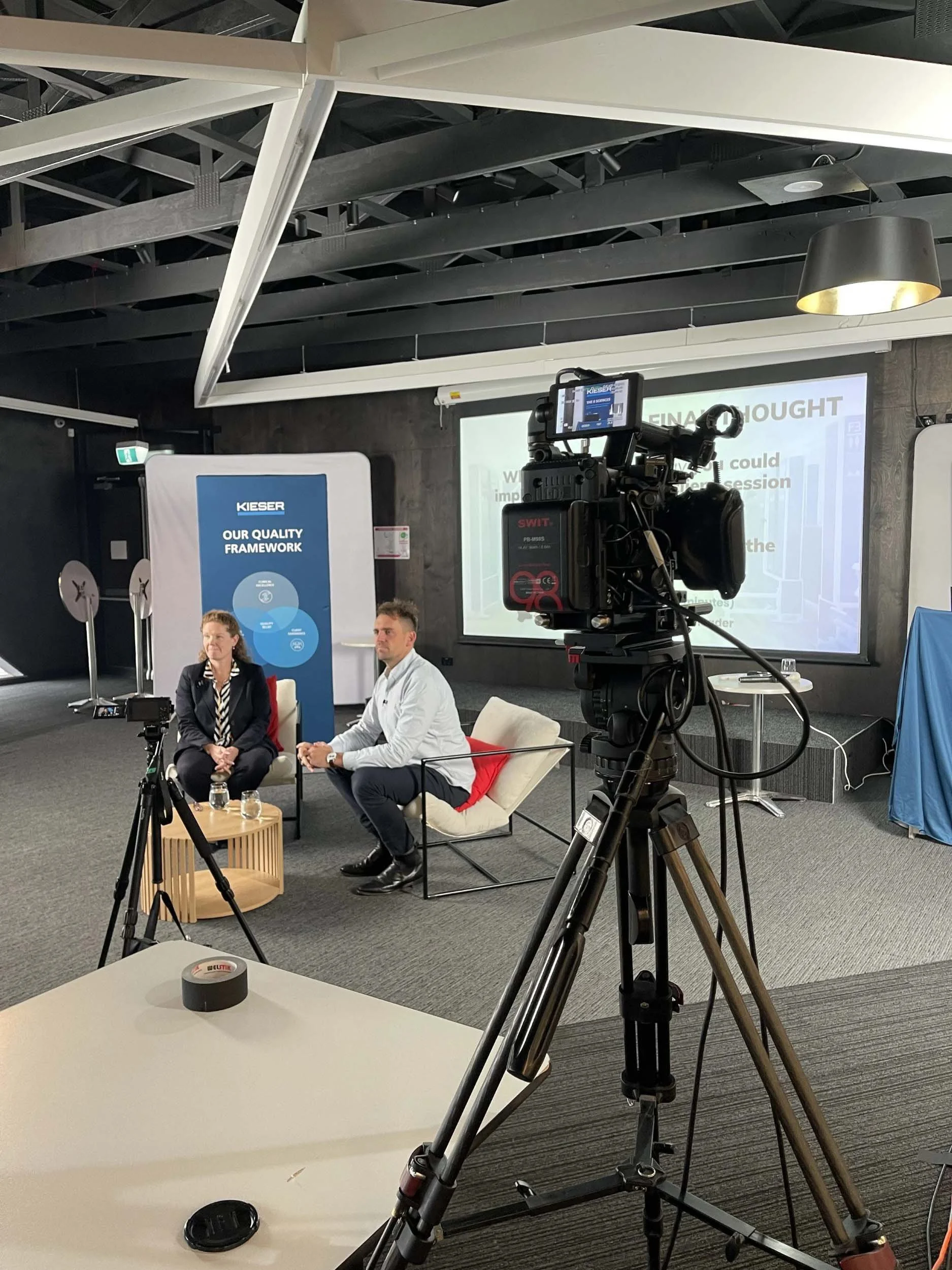 A camera on a tripod capturing a panel discussion with two people seated on chairs in an office or conference room. Behind them is a large screen displaying a presentation. There are various equipment and decor items in the room.