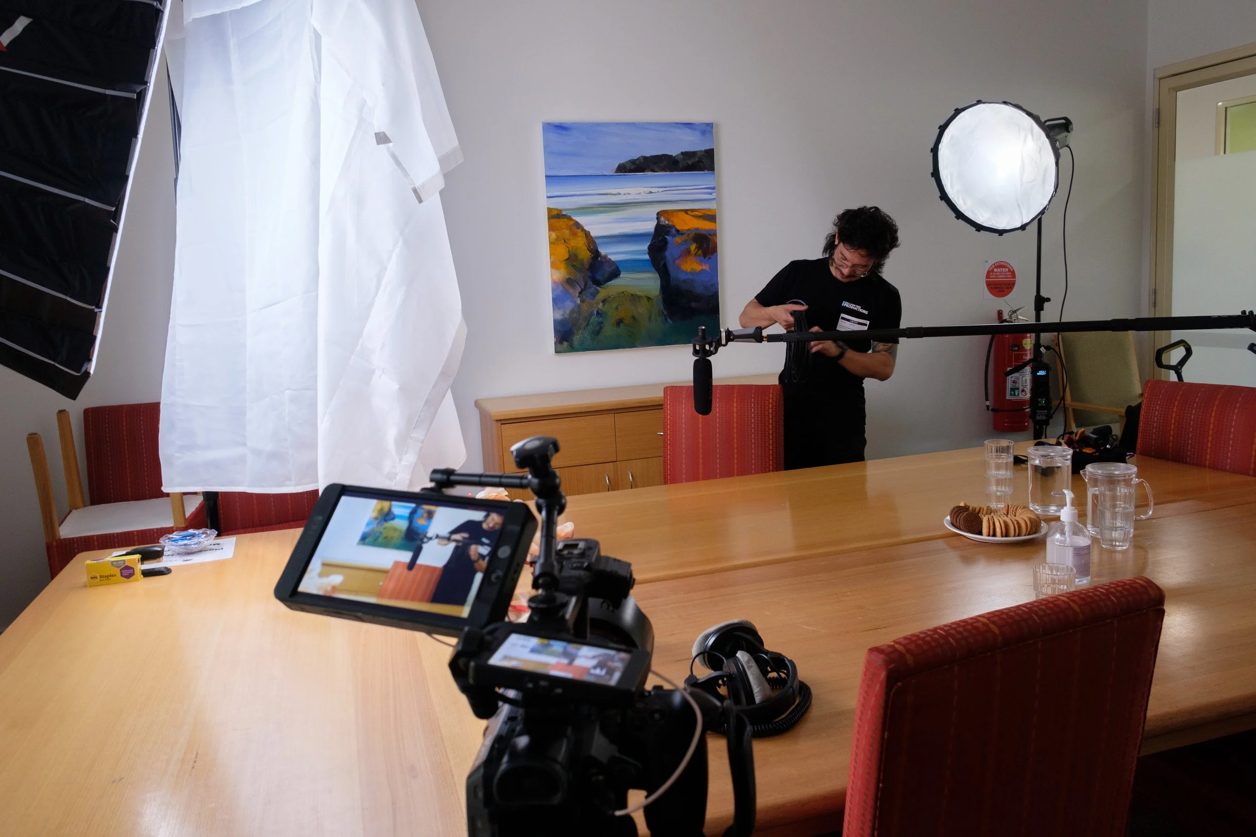 Camera at the bottom of the screen in the foreground with key light on the left and hair light on the right. Ben is adding an XLR cable to the microphone above interviewees chair