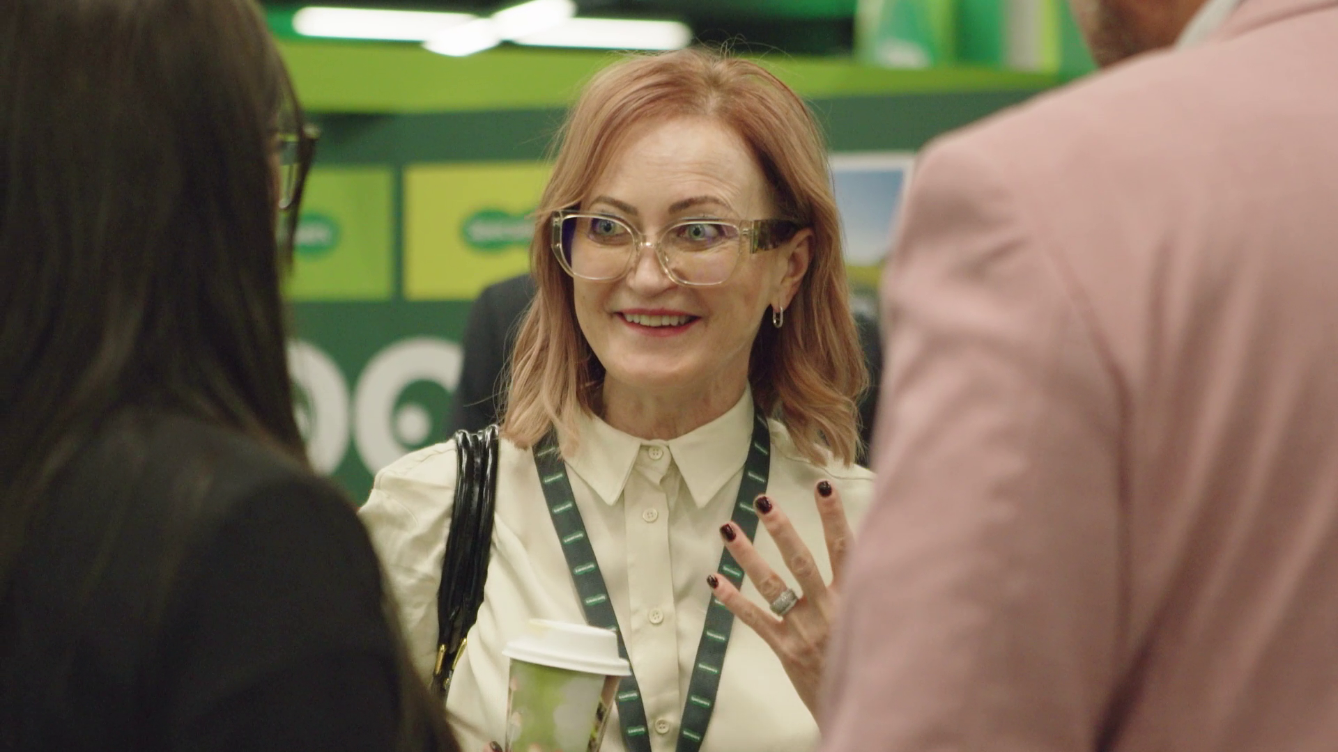 A woman with reddish hair, glasses, and earrings speaking to two people at a store or conference, holding a coffee cup.