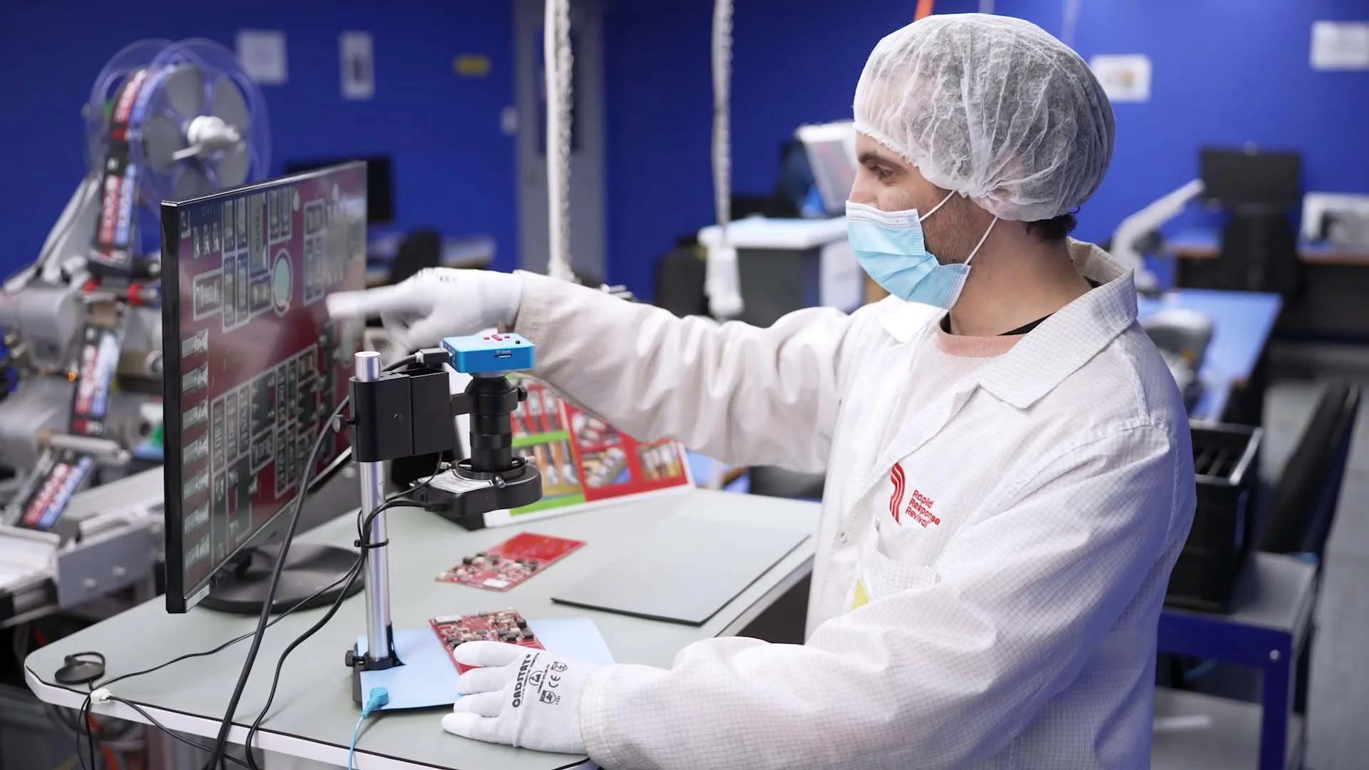 A technician in a lab working on electronic circuit boards at a workstation with a computer monitor, wearing a hairnet, face mask, and gloves.