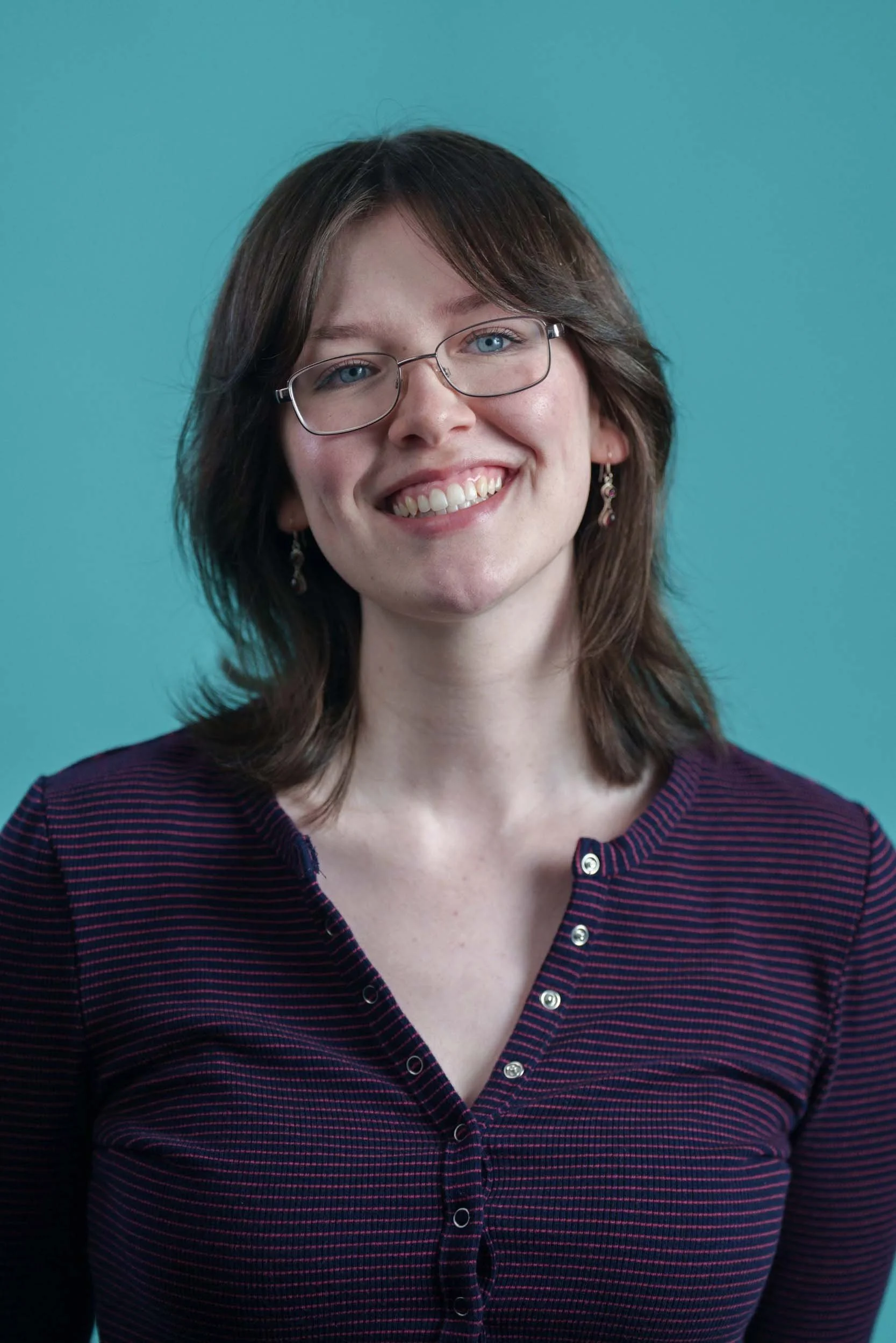A woman with glasses and shoulder-length brown hair smiling, wearing a dark purple and red striped shirt, standing against a teal background.