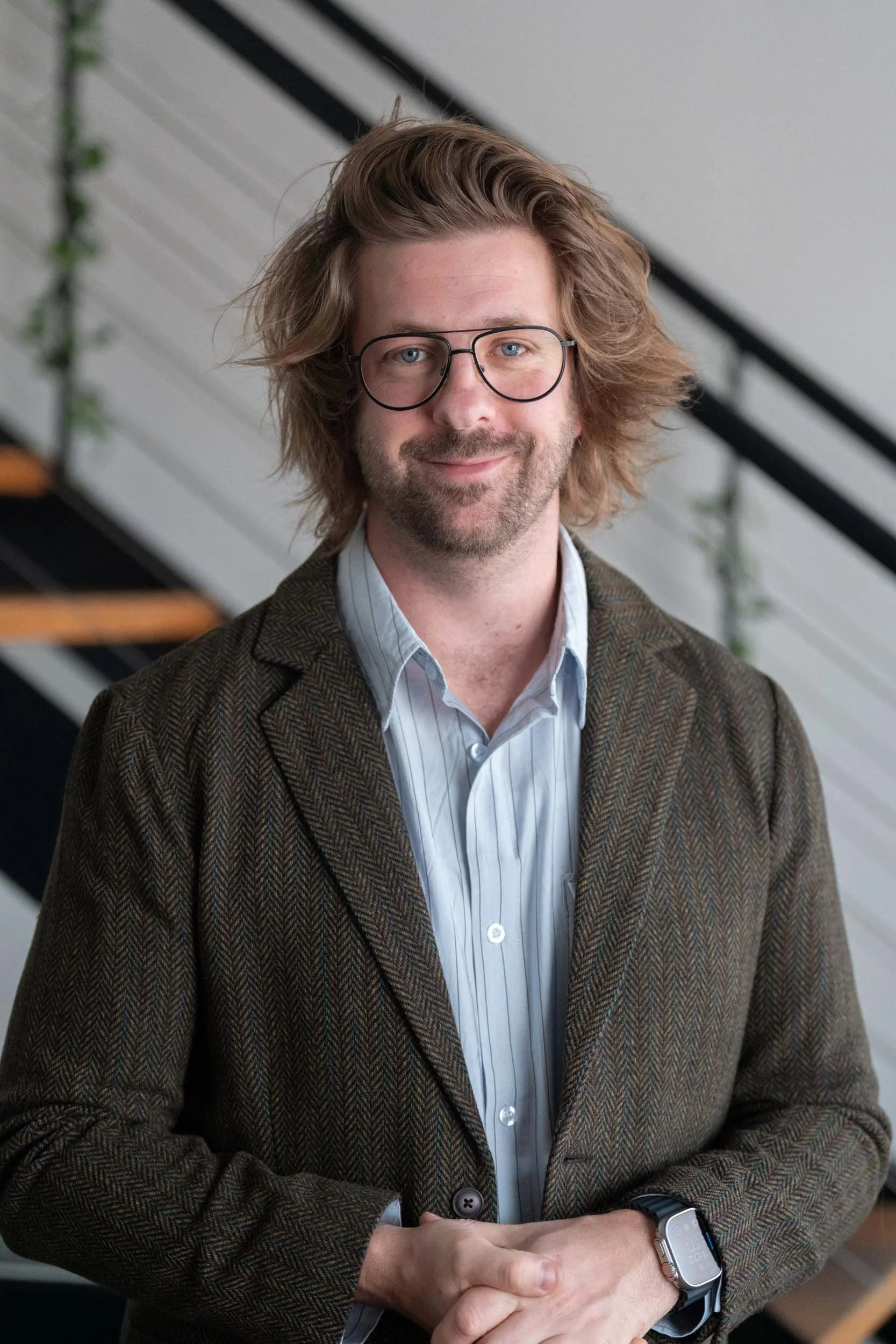 A man with shoulder-length brown hair, glasses, and a beard wearing a plaid blazer over a light blue collared shirt, standing indoors with a staircase and white wall in the background.