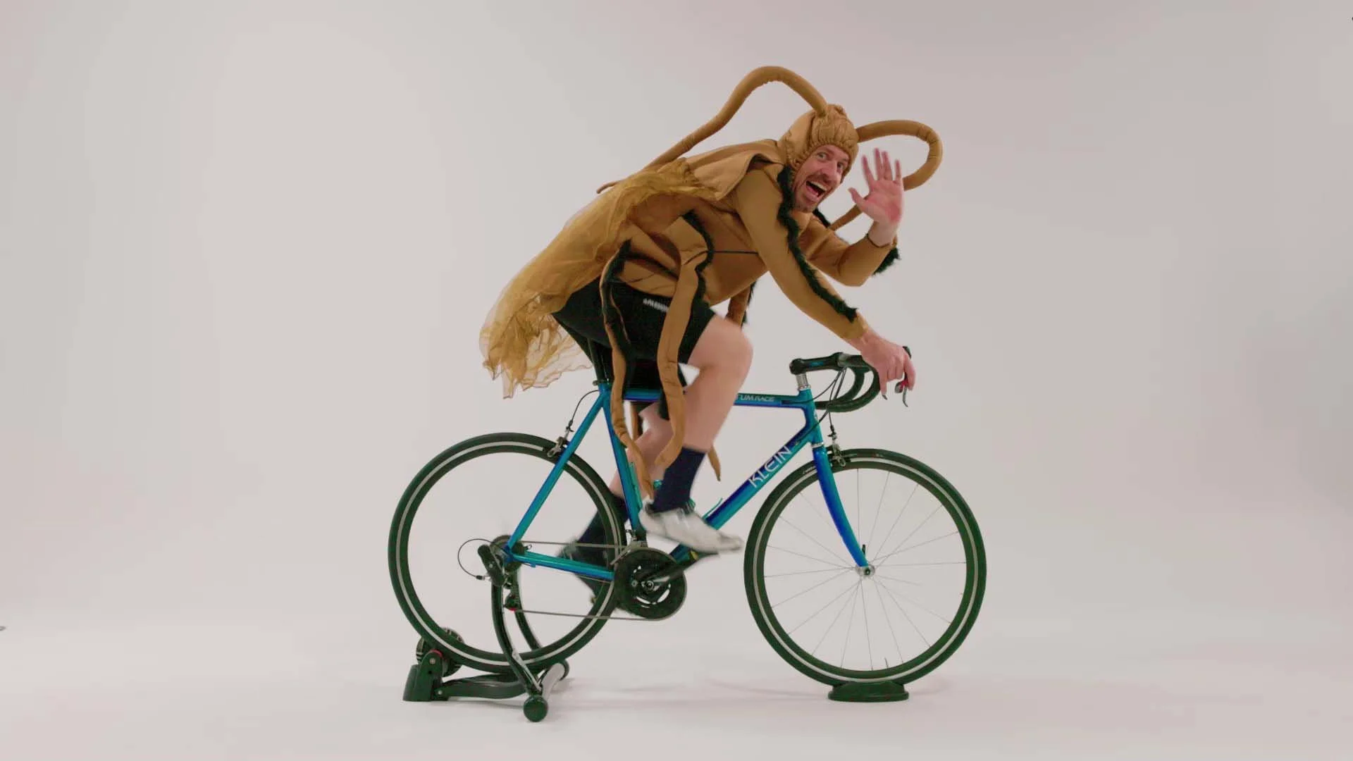 A man dressed in a humorous costume resembling a crab on a blue bicycle, waving and smiling against a plain white background.