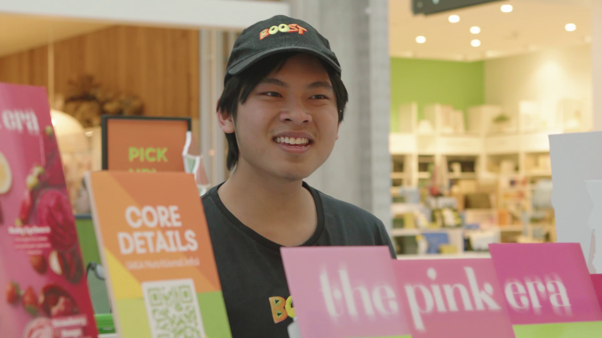 A young man smiling at a Pinkberry frozen yogurt shop, wearing a black cap with a "BOOST" logo and a black t-shirt, standing behind a Pinkberry sign and colorful promotional materials.