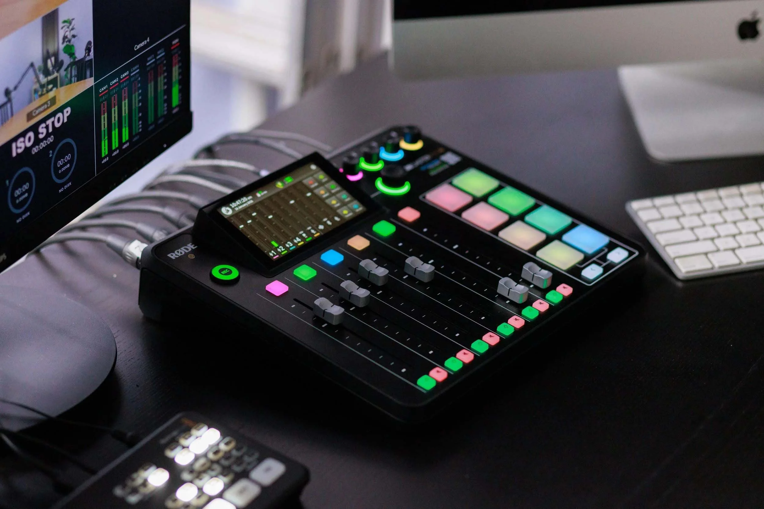 A music production workspace featuring a computer monitor displaying audio tracks and levels, a portable digital audio mixer with colorful illuminated buttons and faders, and a wireless keyboard, all on a dark surface.