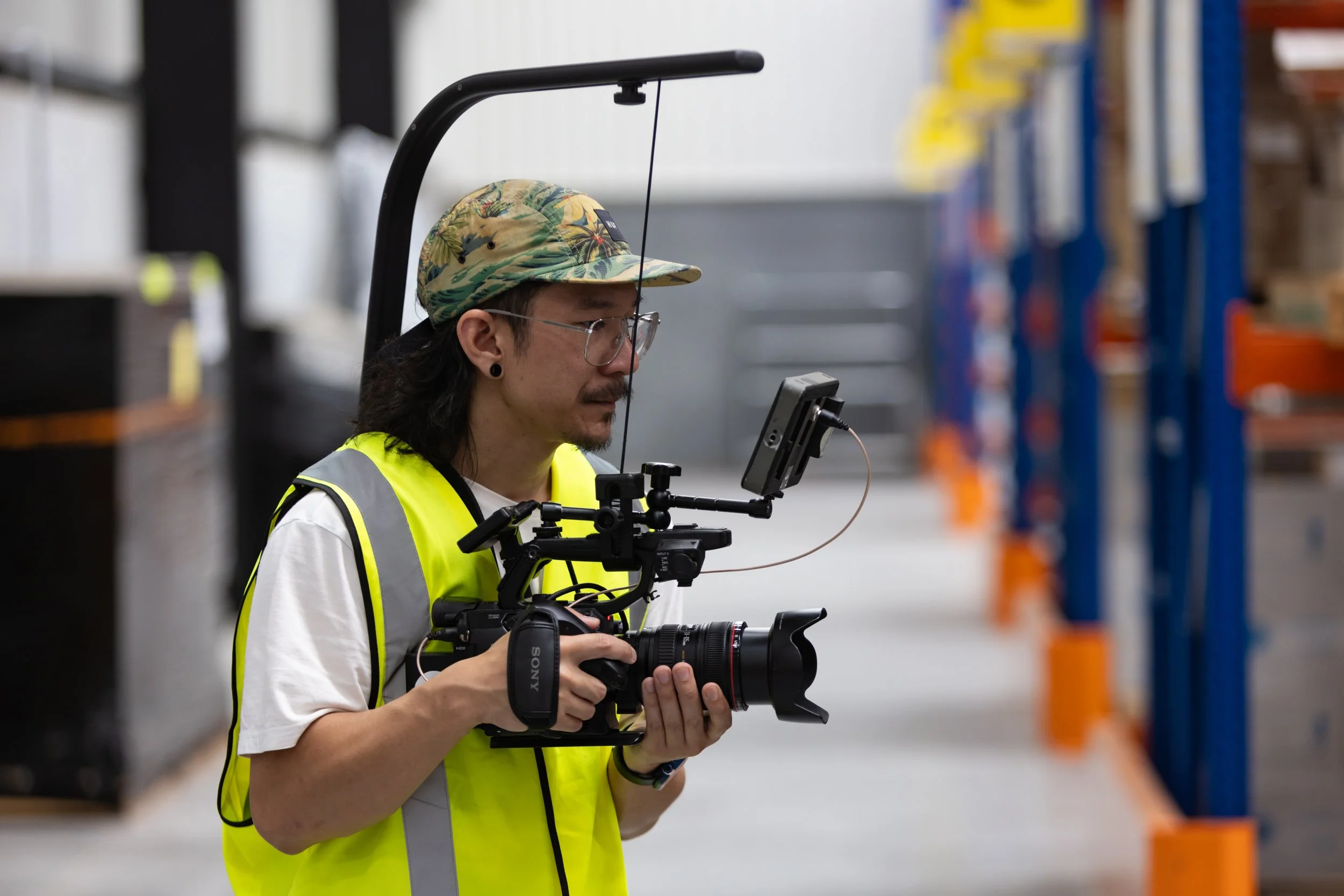 A man wearing a camouflage hat, glasses, a yellow safety vest, and a white shirt records video with a professional camera in a warehouse with blue shelving units and boxes.