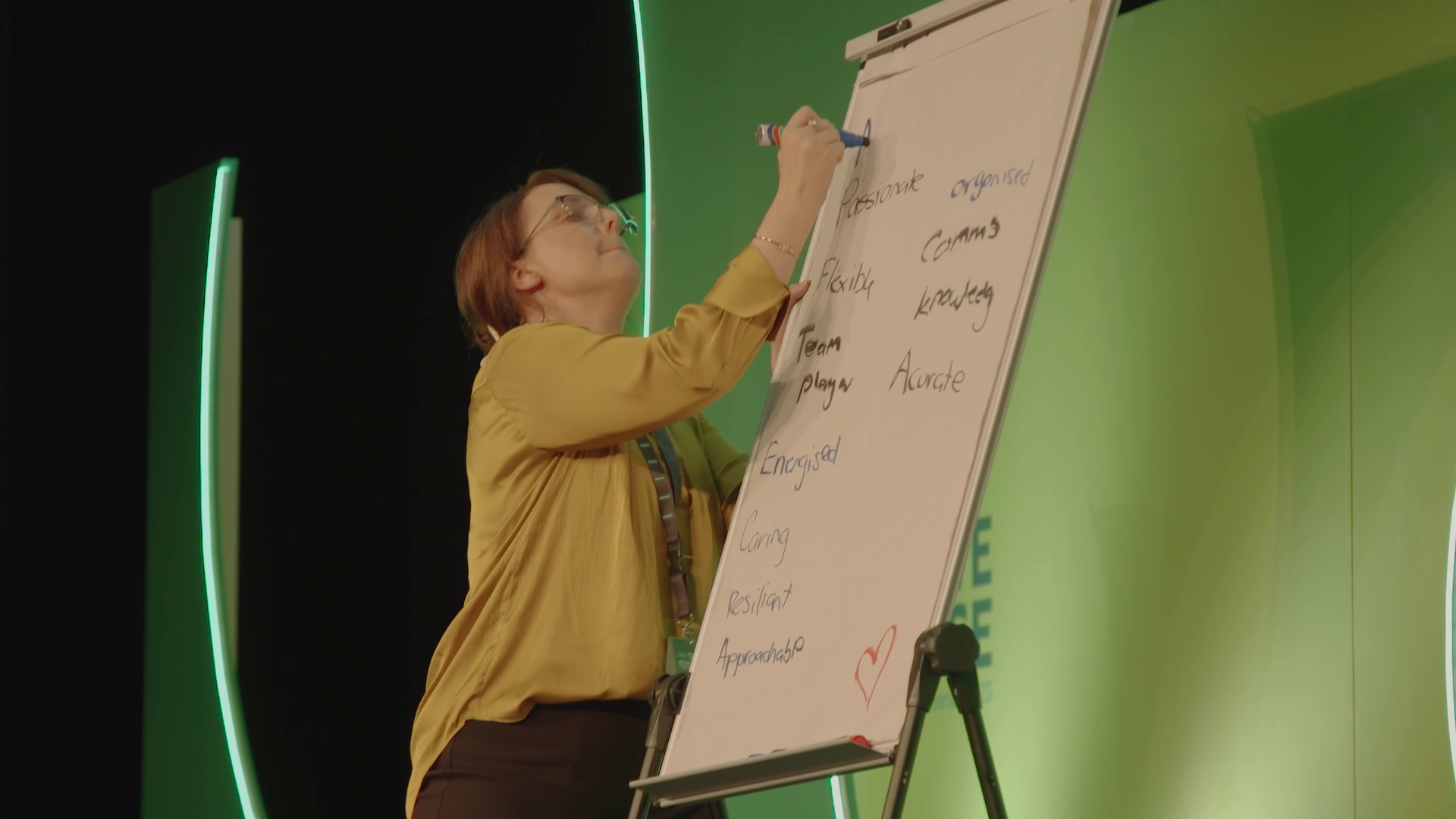 A woman writes on a whiteboard during a presentation or workshop, with notes on skills and qualities like passionate, organized, flexible, knowledge, team, accurate, and heart symbol.