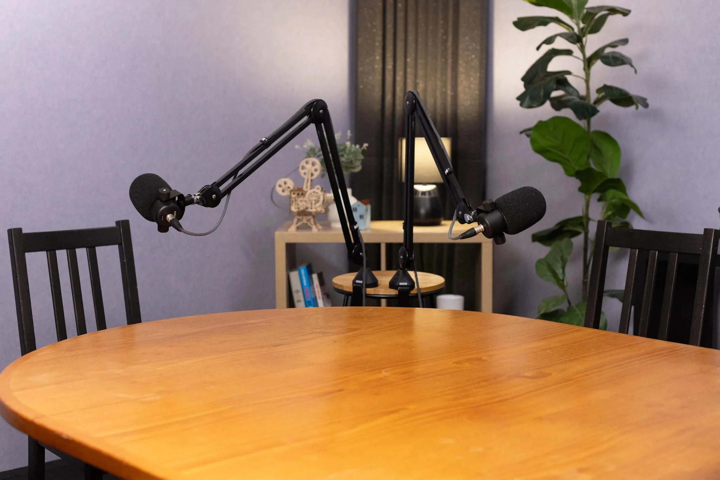 A wooden table in the foreground with black chairs around it. In the background, there are two microphones mounted on adjustable arms, a small bookshelf with books and decorative items, a potted plant, a small wooden rack, and a lamp against a purple