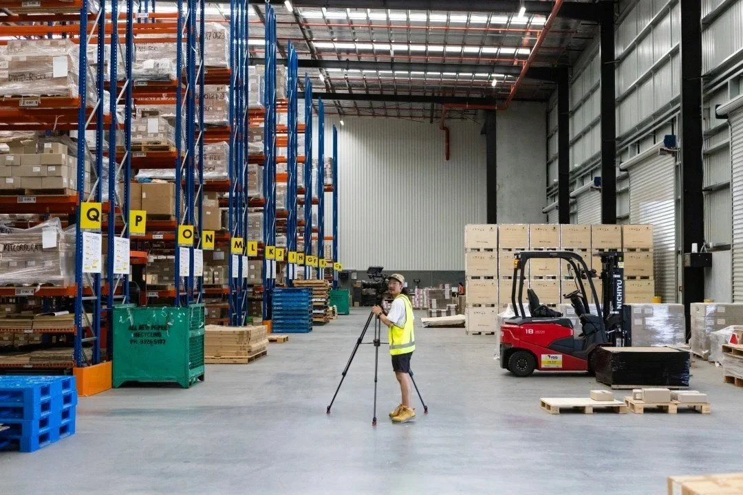 A warehouse with tall blue and orange shelving units filled with boxes. A worker wearing a yellow safety vest, black shorts, and a cap is operating a tripod-mounted camera. There are a red forklift and pallets with boxes on the concrete floor.