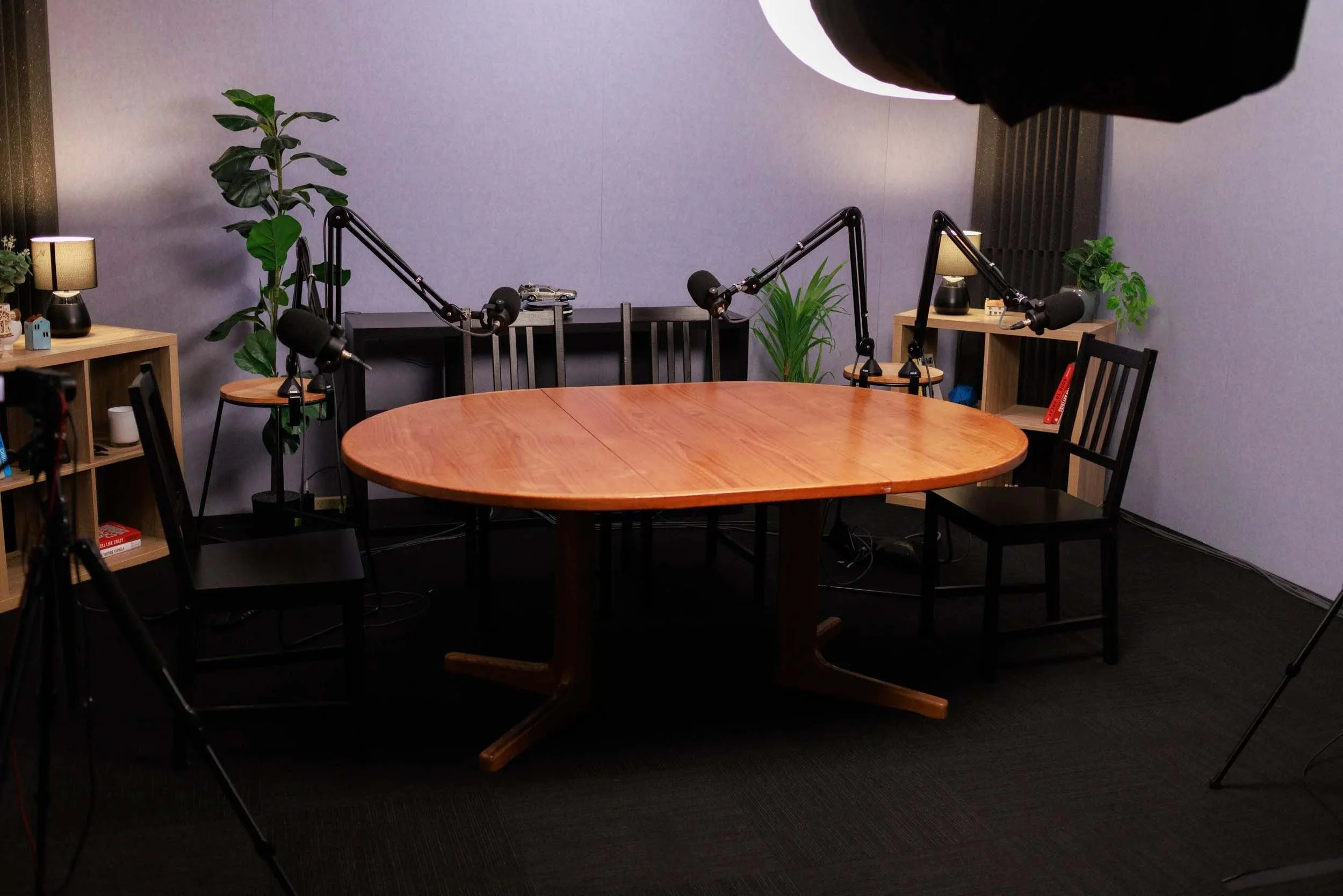 A podcast recording studio with a round wooden table, black chairs, and three microphones on adjustable arms. There are two side tables with lamps, plants, and books, set against a gray wall with black accents.