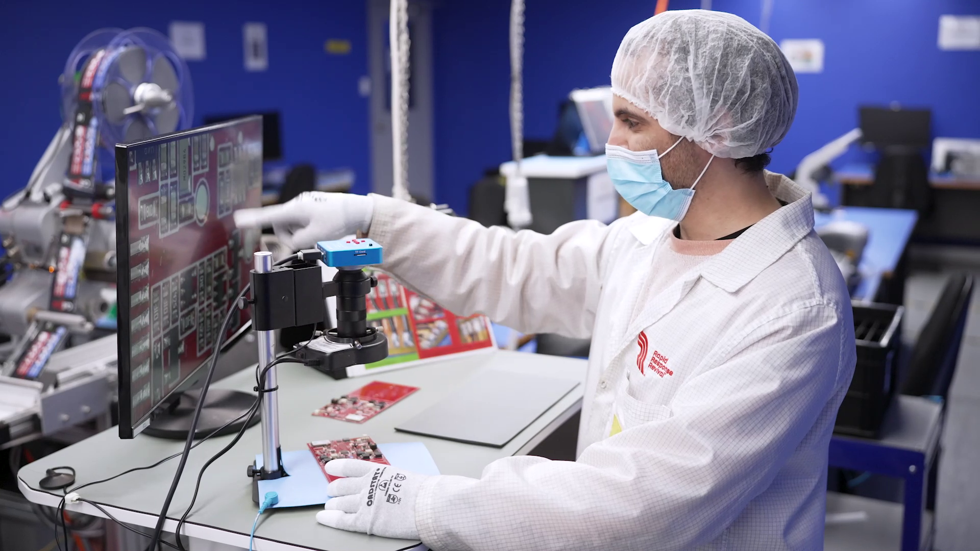 A worker in a cleanroom working on electronics with a computer monitor, wearing a hairnet, face mask, gloves, and a white lab coat in a high-tech manufacturing lab.