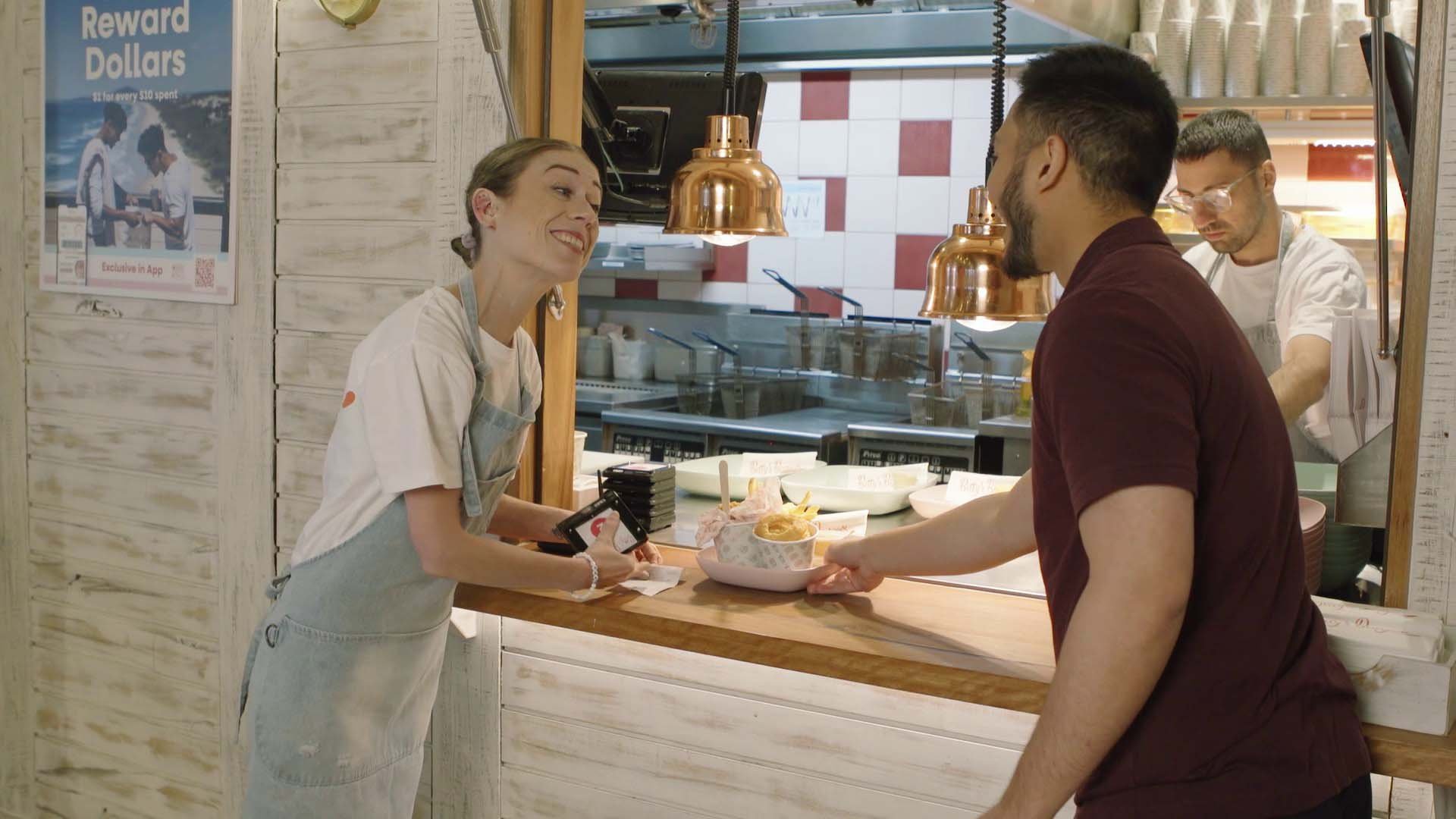 A woman at a restaurant counter receiving food from a man. There are two workers in the background, one wearing glasses and a white apron. The setting appears to be a casual eatery with a white rustic decor and copper pendant lights.