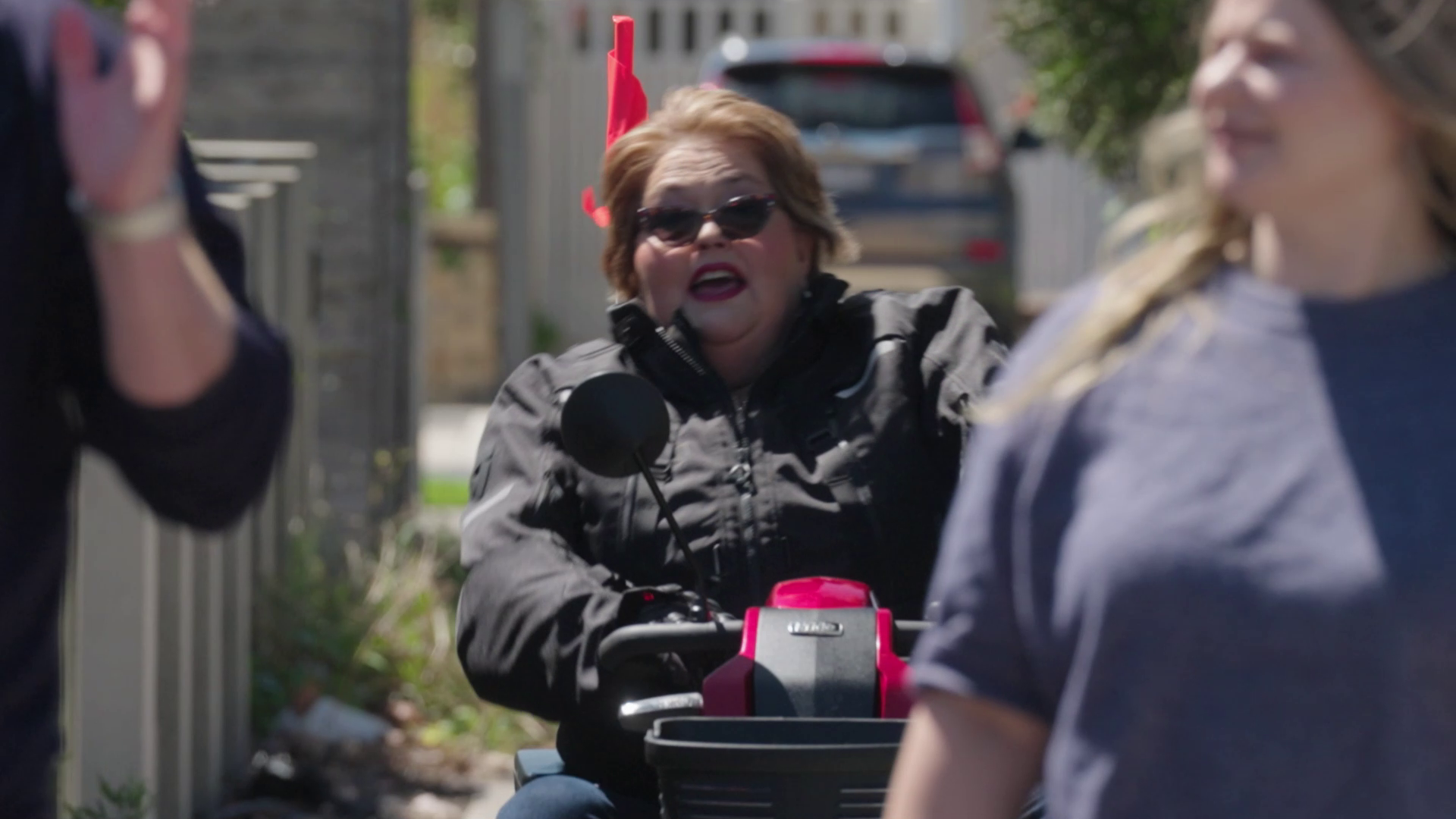 A woman with short brown hair wearing sunglasses and a black jacket riding a pink and black mobility scooter on a sidewalk.