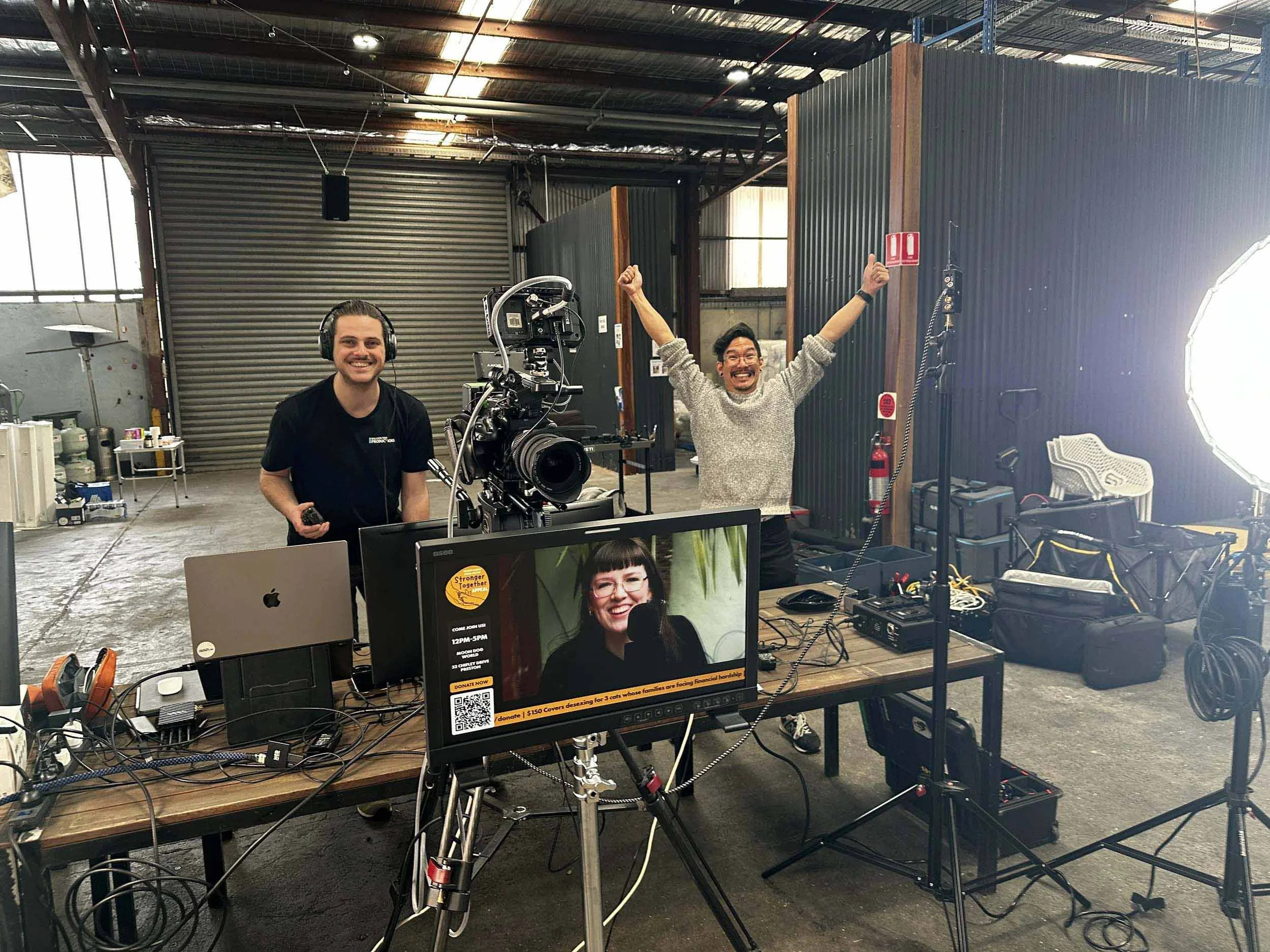 People in a warehouse or studio setting with camera equipment, two individuals smiling and raising their arms, computer monitors, and various electronic devices on the table.