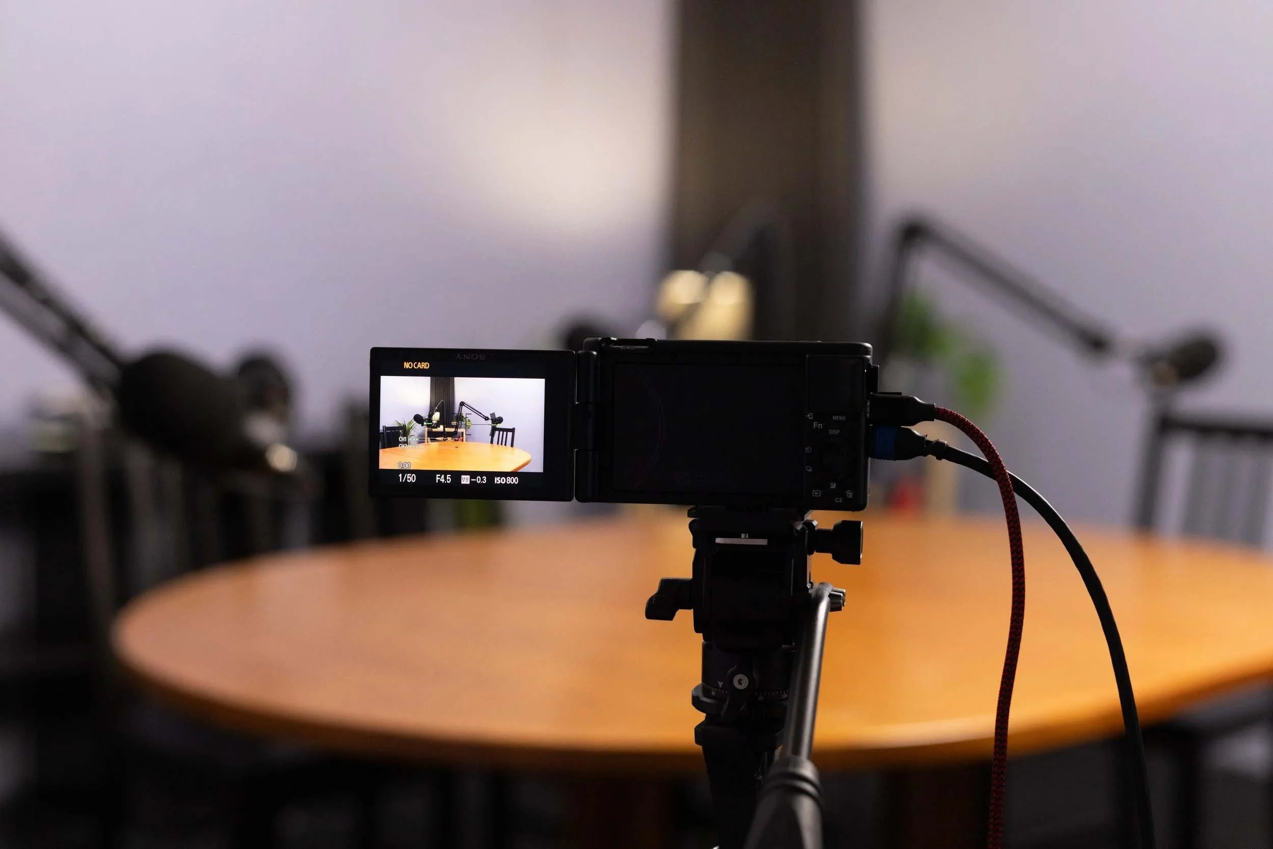 Camera on a tripod capturing a table around which multiple chairs are arranged, with a plain wall in the background.