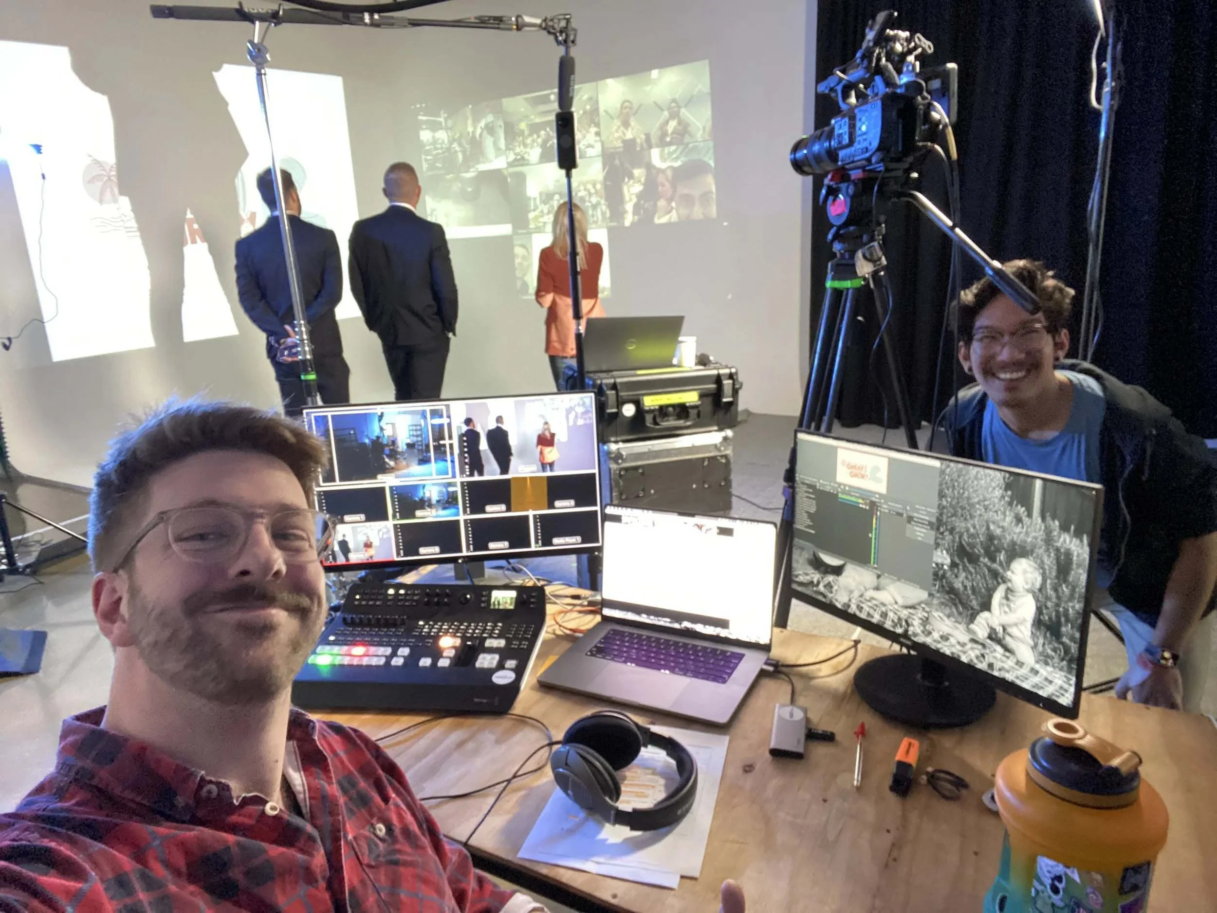 A group of people in a production studio. Two individuals are on a stage in front of a large screen showing a collage of images and others wearing suits. There are people at desks with computers and a person smiling at the camera in the foreground.