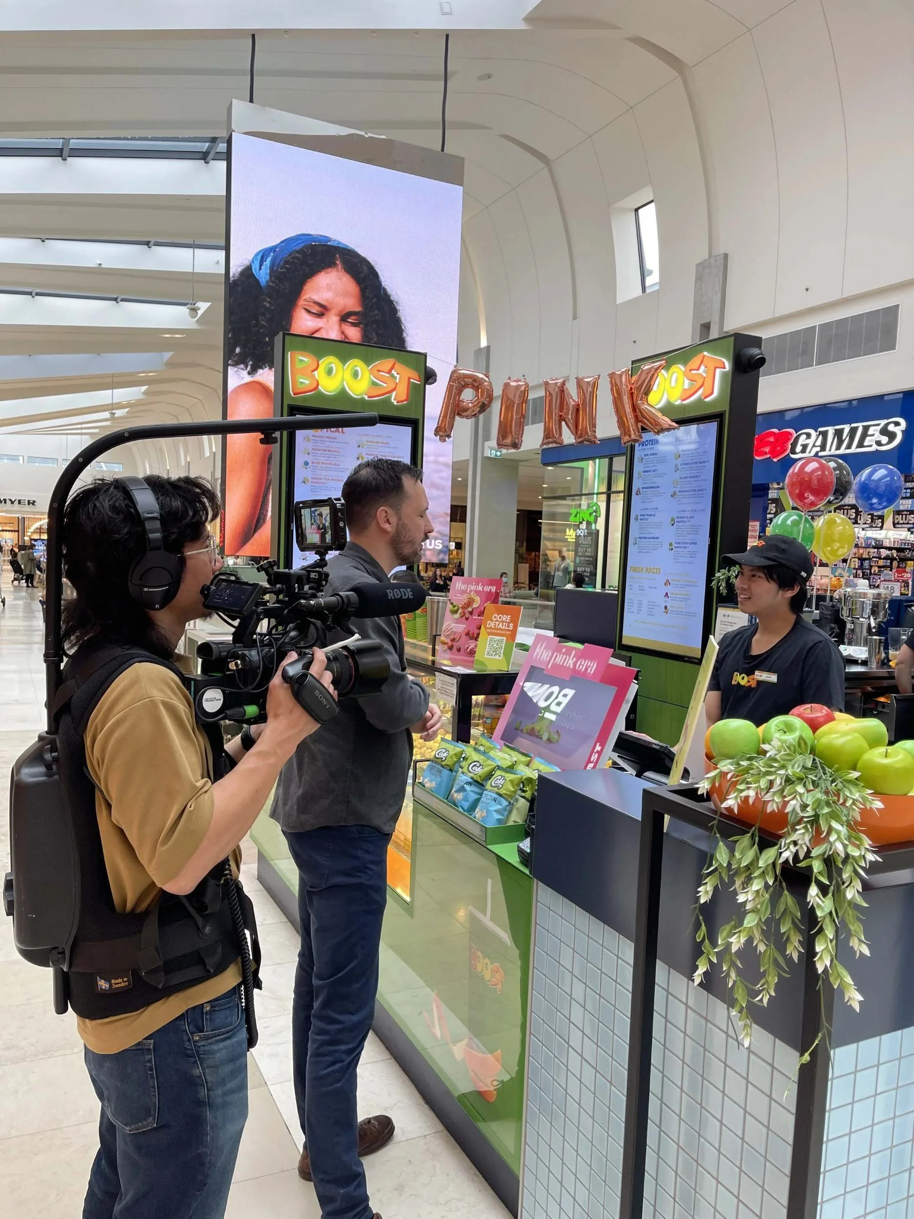 People placing orders at a juice bar inside a shopping mall, with balloons and a large advertising screen in the background.