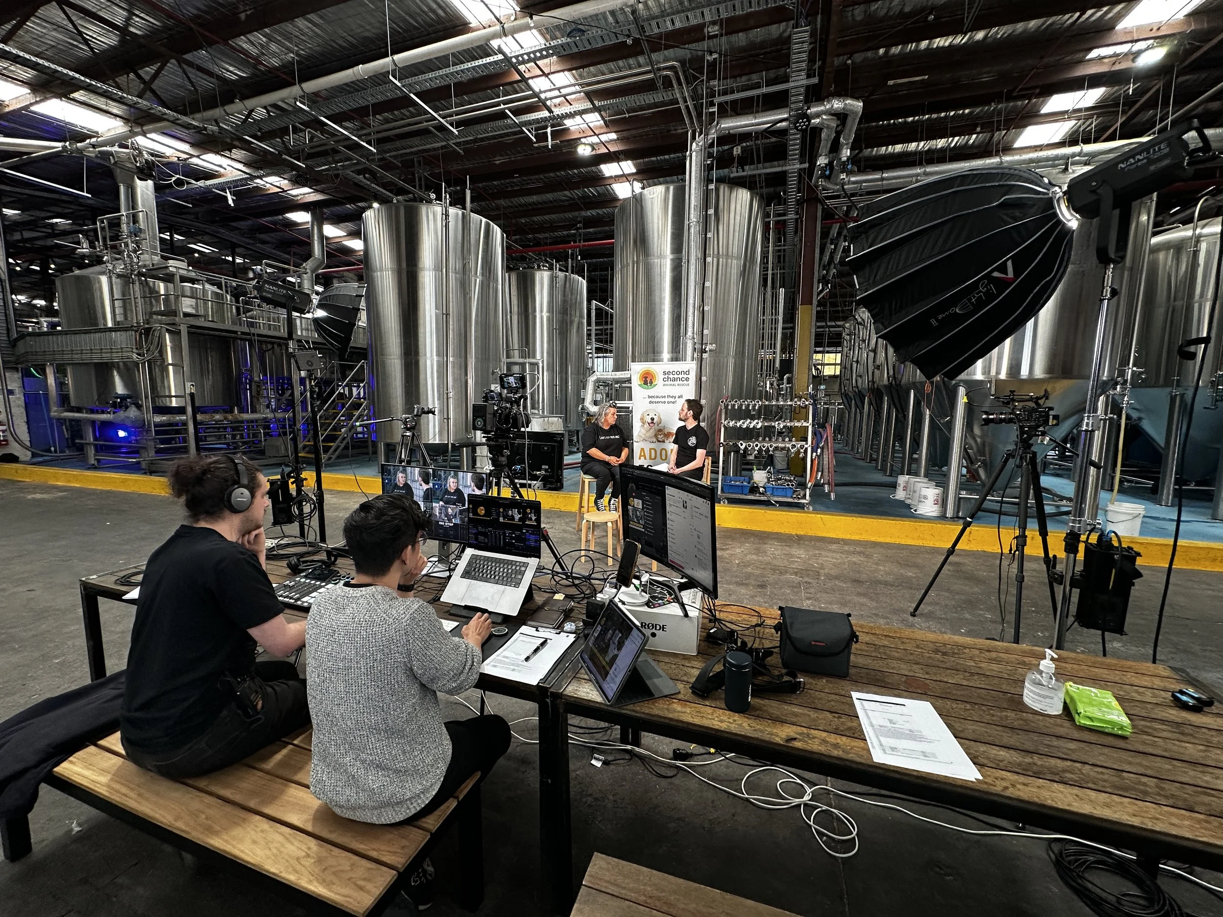 A behind-the-scenes view of a video production setup inside a brewery, with two people being interviewed on a small stage and three technicians working at a table with various electronic equipment and monitors.