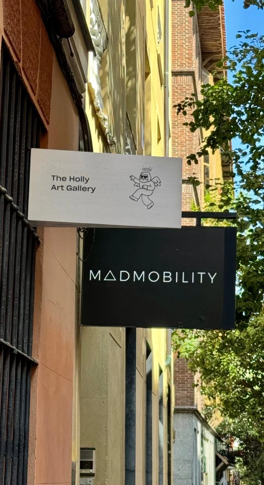 Signs for The Holly Art Gallery hanging on a building exterior, with trees and a brick building in the background.