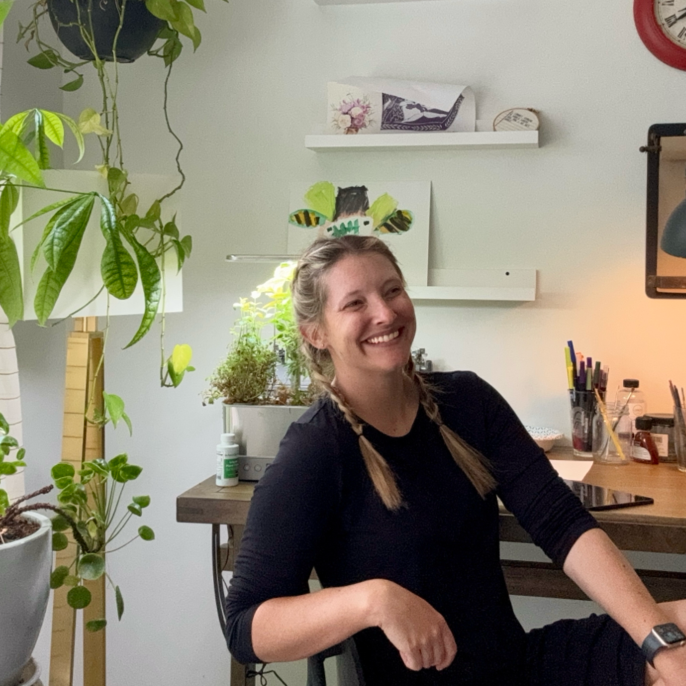 Smiling woman with blonde hair in braids sitting at a desk surrounded by plants in a cozy, well-lit room.