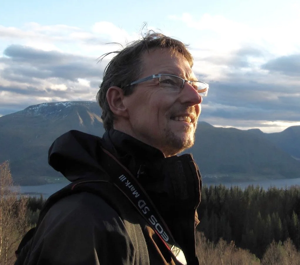 Man with glasses smiling outdoors during sunset, with mountains, a lake, and a cloudy sky in the background.