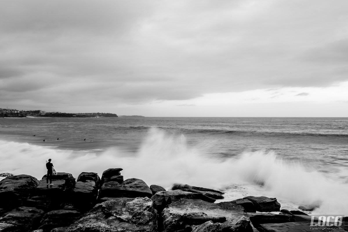 A surfer looks out at water as the waves crash against the rocks at Manly Beach in Australia.
