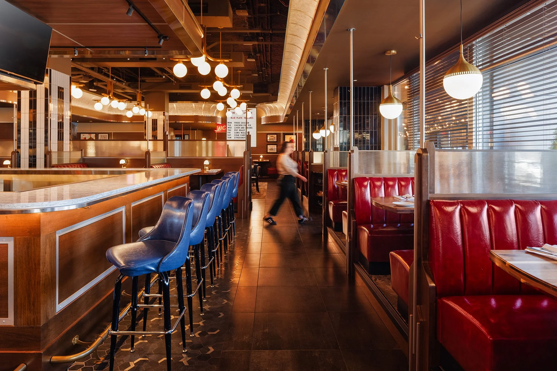 Interior of a modern restaurant with a bar on the left, red booths on the right, large spherical pendant lights hanging from the ceiling, wooden accents, and a waiting staff member walking through the space.