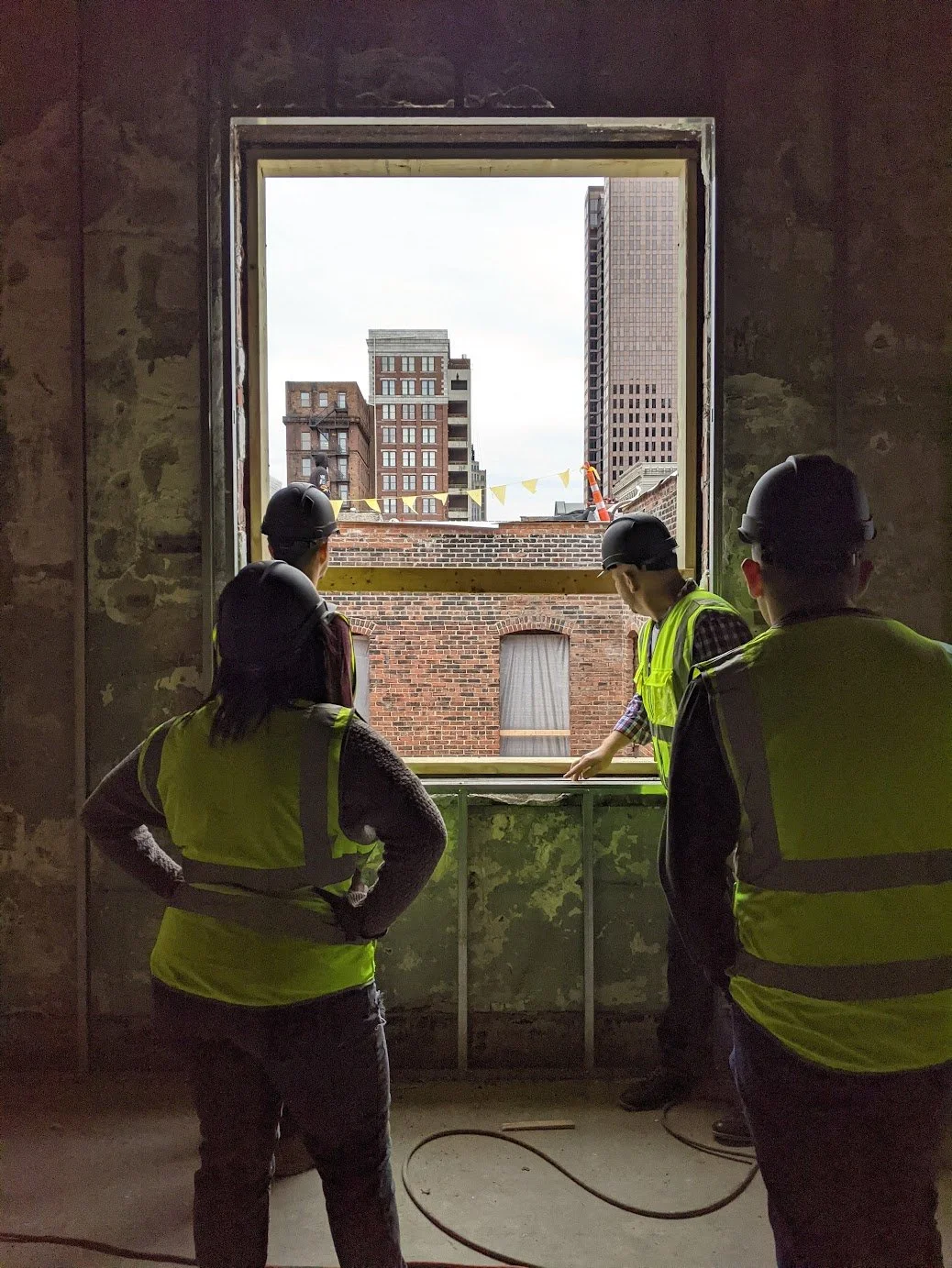 Four construction workers wearing yellow safety vests and helmets looking out an open window into an urban cityscape with buildings of various heights.