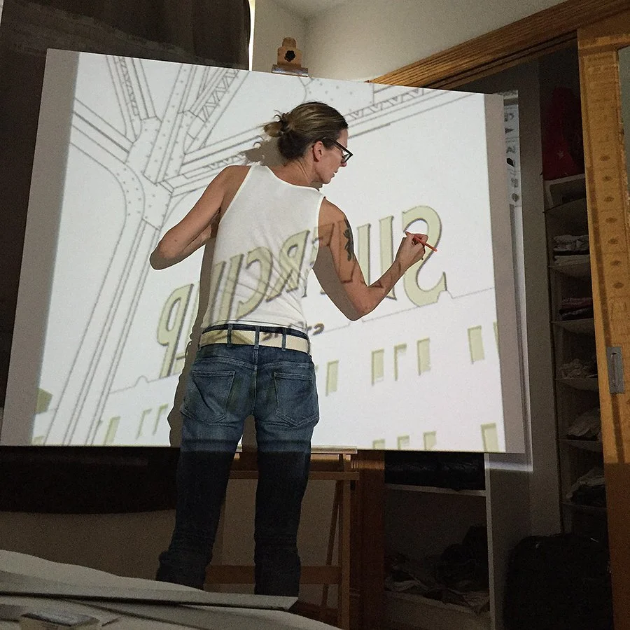 Person with long hair and glasses writing on a large screen or whiteboard, with the word 'Sincerely' visible. The person is wearing a sleeveless white top and blue jeans, and is standing in a room with wooden shelves and a cabinet.