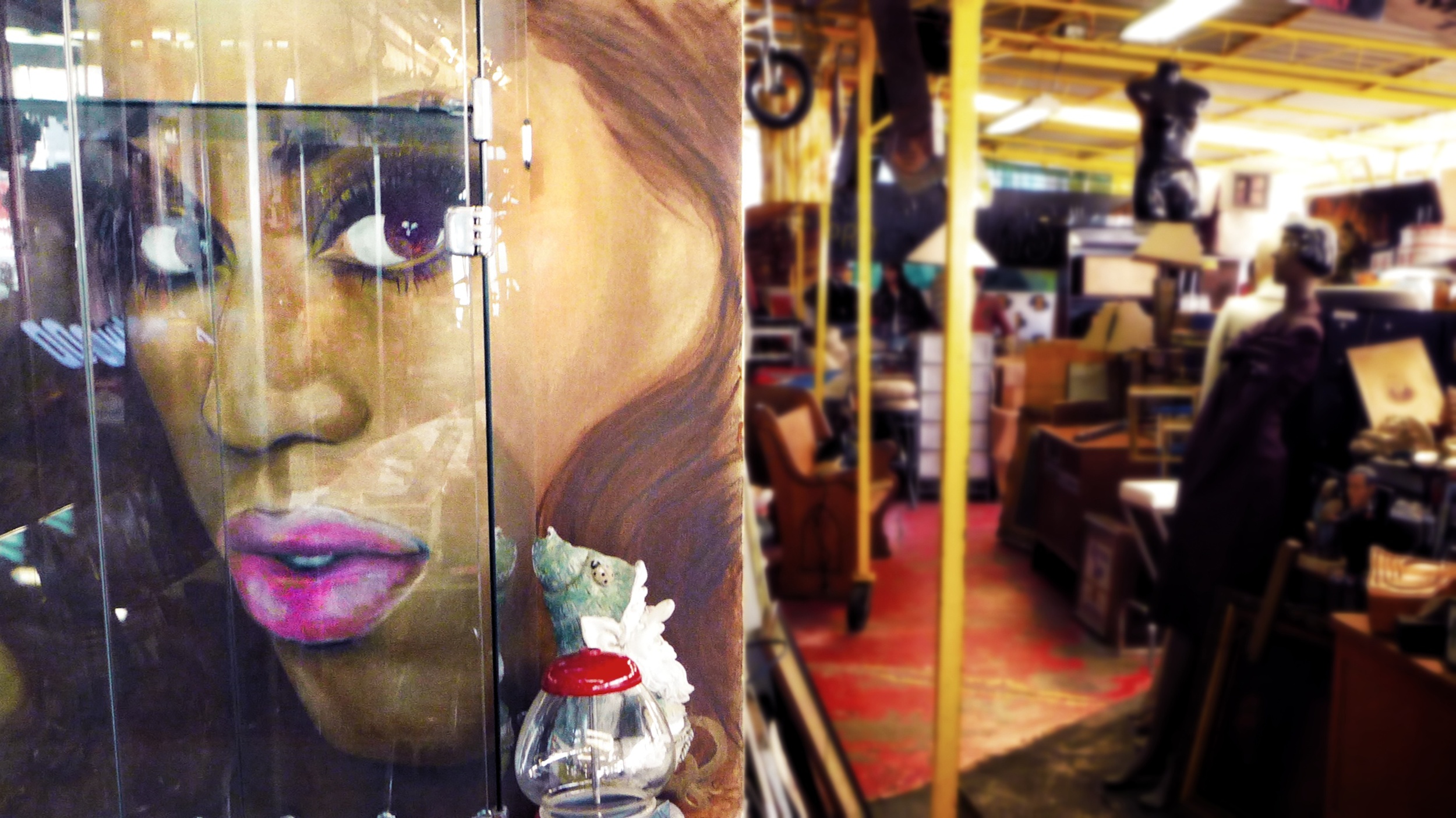 Close-up of a painted face with striking eyes and bright pink lips, on a glass display case in a store or gallery setting.