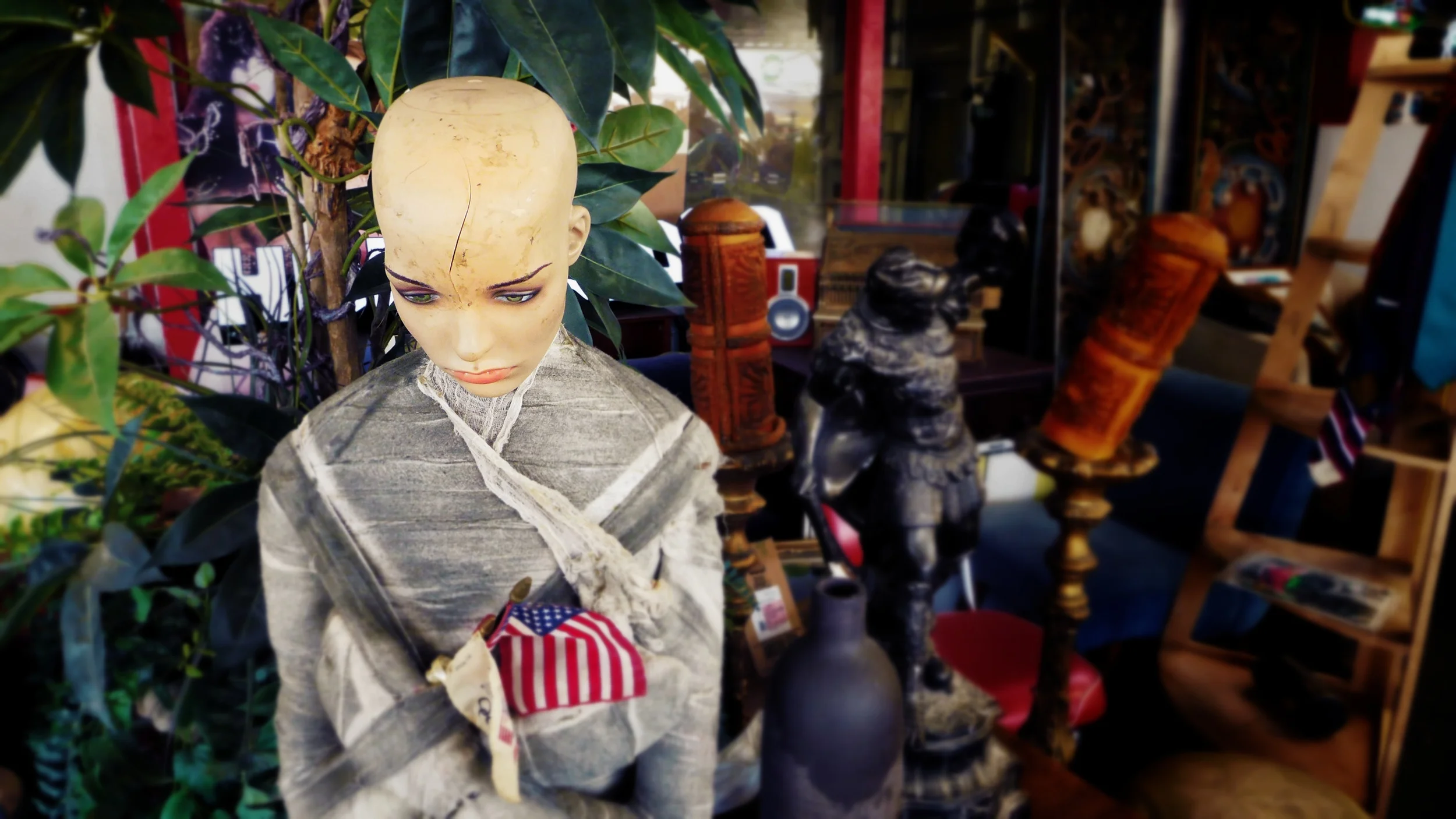 A mannequin with a bald head and painted facial features wearing a gray outfit, holding a small American flag, surrounded by various decorative objects and plants in an indoor setting.