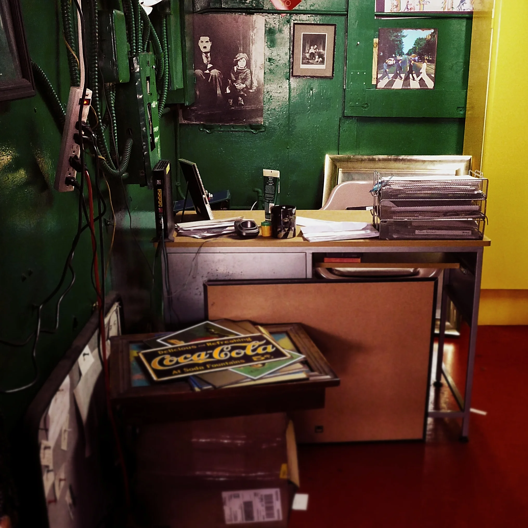 A cluttered desk with scattered papers, a magazine with a Coca-Cola advertisement, and a wire organizer. In the background, there are framed vintage photographs and a crosswalk scene on the wall. The room has green and yellow painted walls.