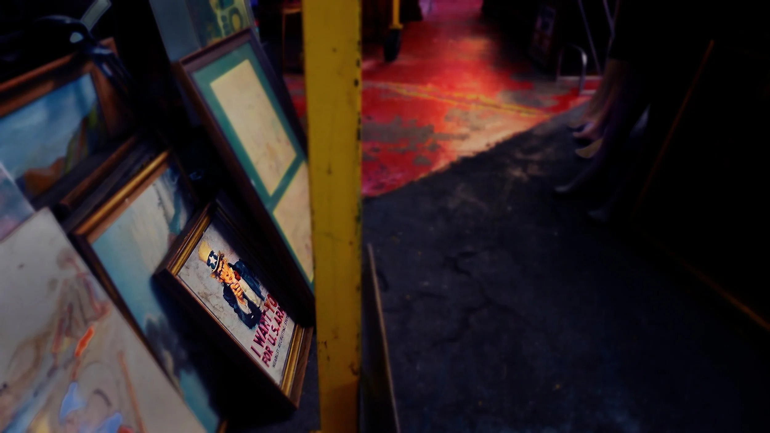 A view of a cluttered space with framed pictures, one featuring a child in a costume, leaning against a yellow post, with a red and black patterned floor and shadowy background.