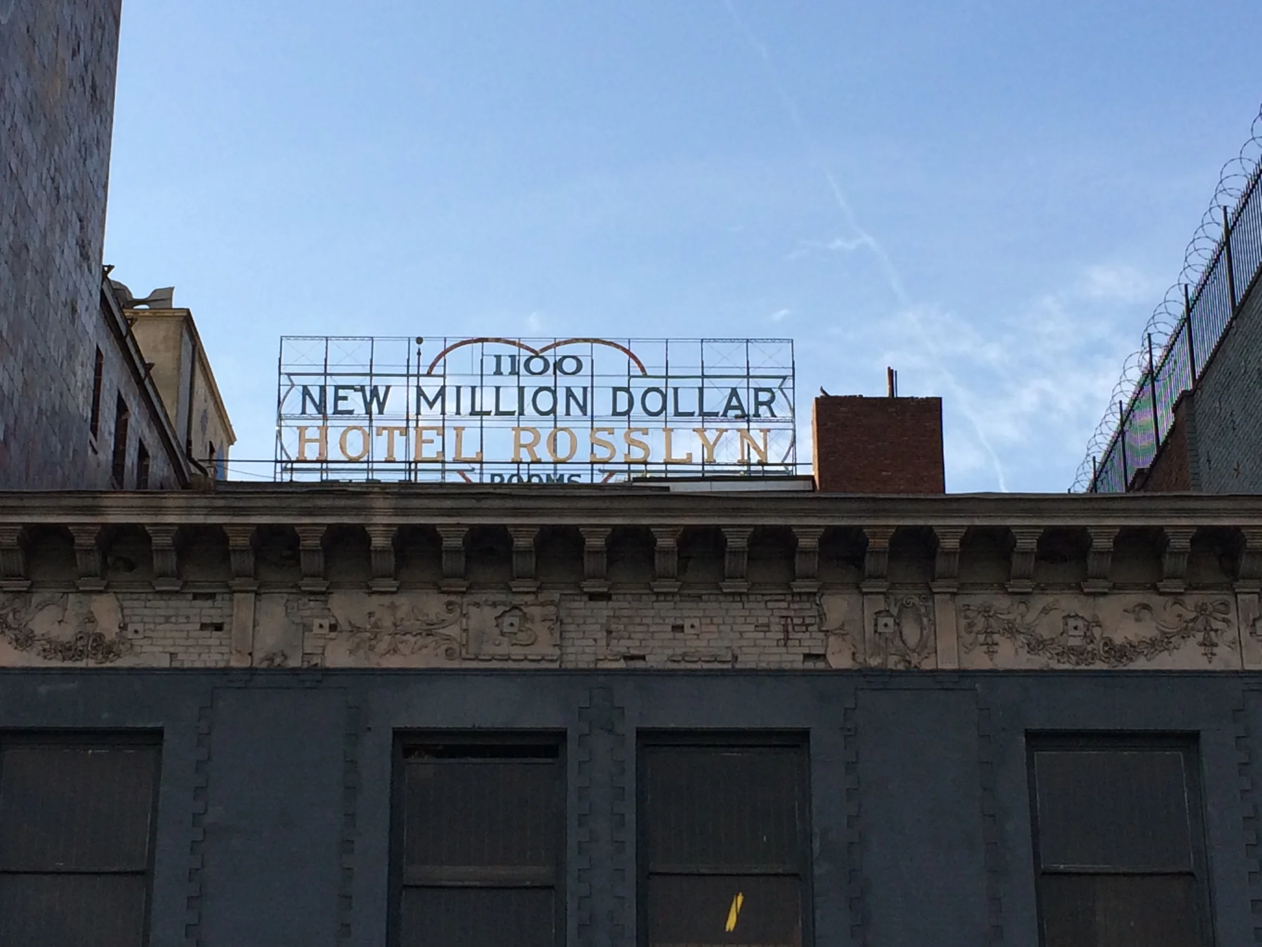 Upper part of a building with a cityscape behind. There is a sign on top of the building that reads 'New Millen Dollar Hotel Rosslyn' with some additional text that is partially obstructed. The sky is mostly clear with some clouds.